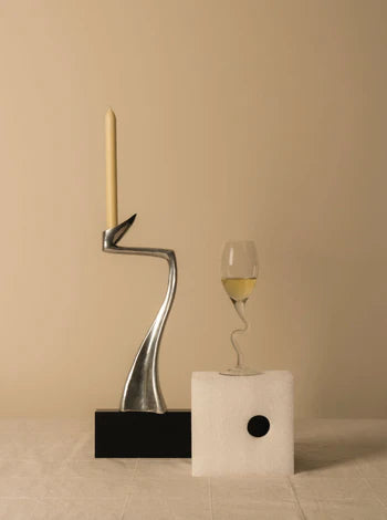 A modern, silver candle holder with a lit beige candle stands on a black base. Next to it is a wine glass with white wine, positioned on a white rectangular block with a circular hole in the center. The background is beige.