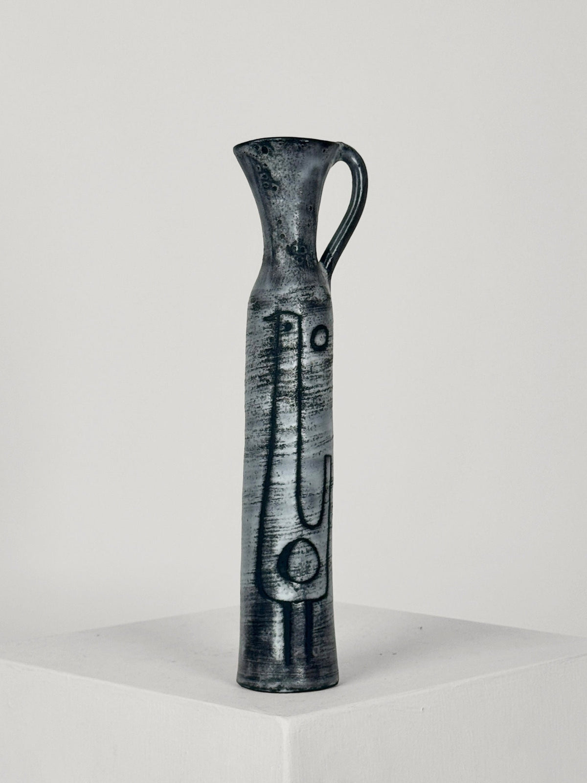 A tall, slender Ceramic Bottle by Jacques Pouchain from Figuresdesign features a narrow neck and a small handle on its upper part, adorned with minimalist abstract patterns etched into its dark ceramic surface. The bottle is displayed on a plain white pedestal against a neutral background.