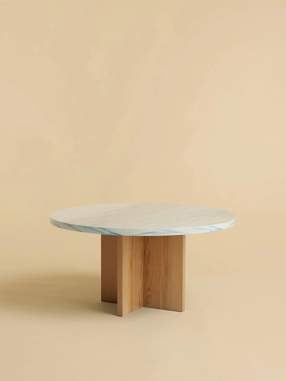 The "Tinian" Coffee Table by Lebanto features a round marble top with a white and blue pattern, artisanally crafted, resting on an oak wood cross-shaped base, all set against a plain beige background.
