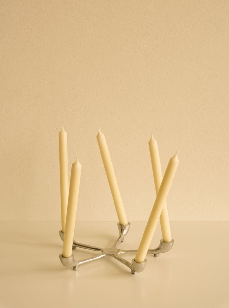 Five beige candles of varying heights arranged in a Les Objets Amsterdam sleek candelabra against a soft beige background.