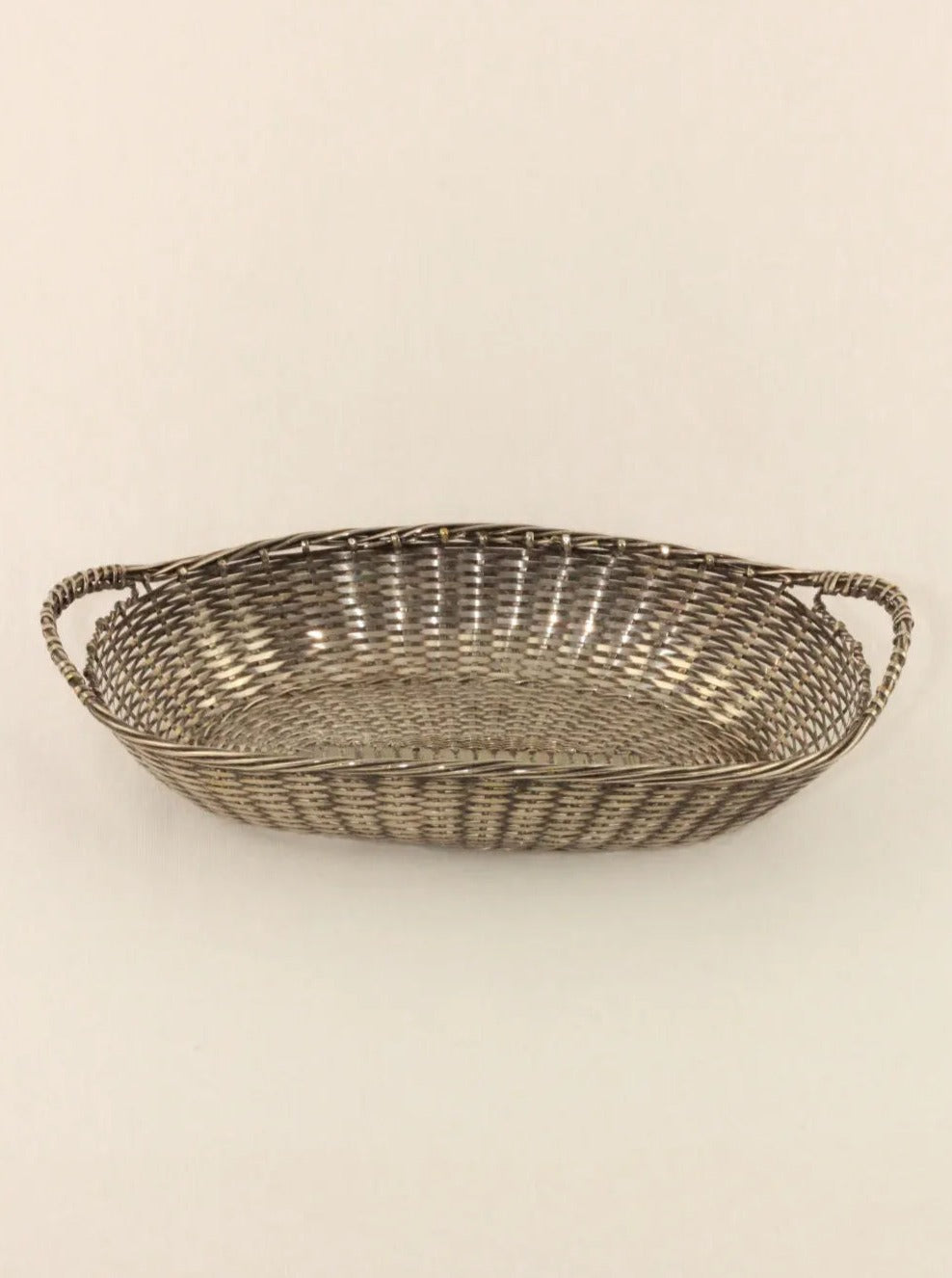A shallow, oval-shaped hand-woven basket with braided handles on either side is displayed against a plain beige background. The Woven Basket by Boga Avante Shop, woven in a tight, uniform geometric pattern, exudes simplicity and elegance.