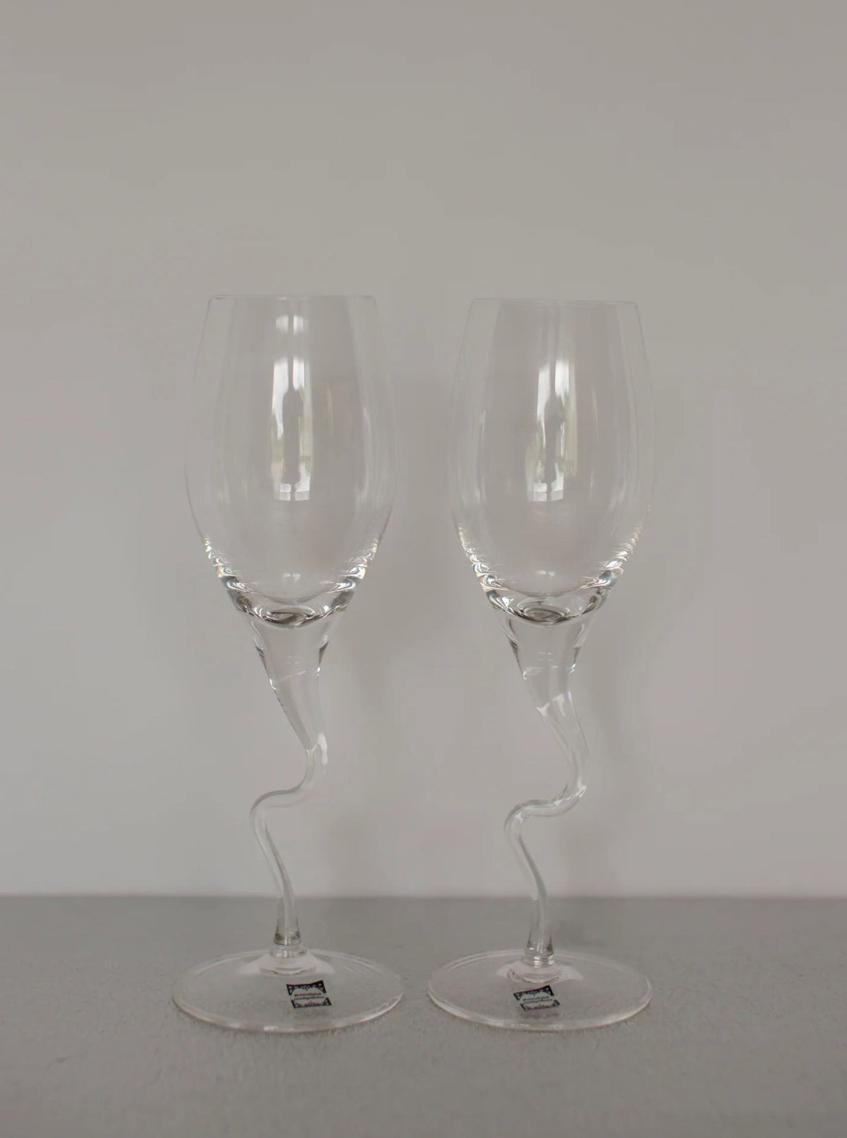 Two clear, intricately designed Set of Vintage Handmade Swirl Champagne Flutes by CU-RA-TED with twisted stems are placed side by side on a gray surface. The artisan-made, artistic shape of the stems adds a vintage charm and sophisticated touch to the otherwise classic glassware.