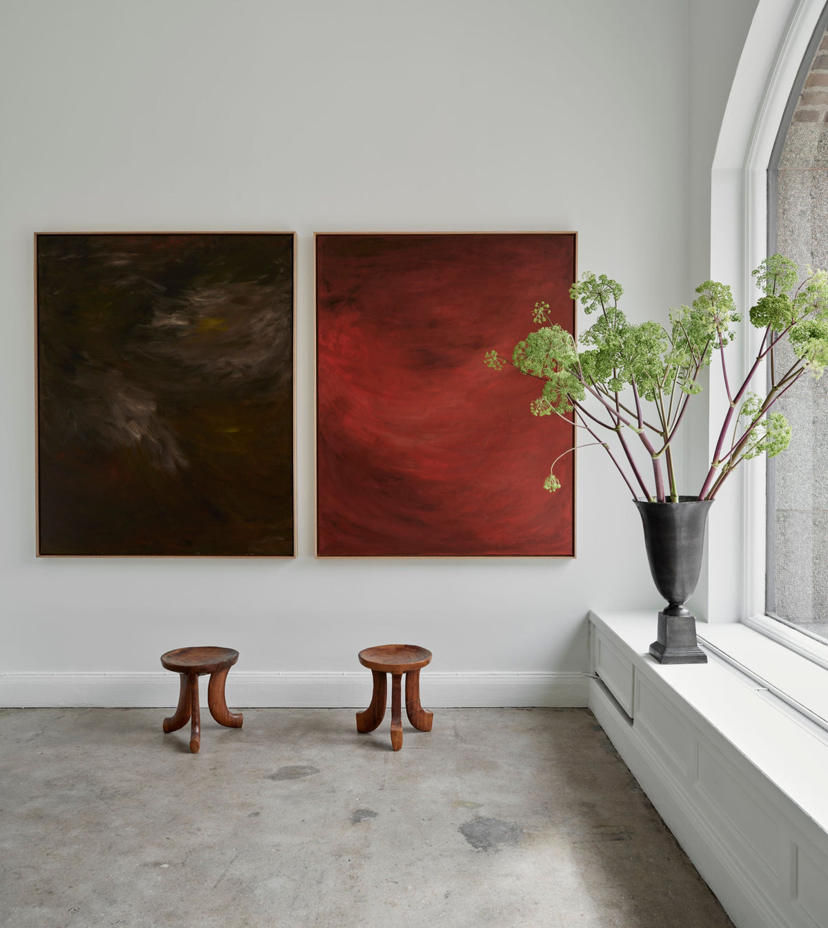 A minimalist gallery room features two abstract paintings from the Terra Incognita (Diptych) - Mixed media on canvas by Mattias Storm—one dark and the other in rich red hues—hanging on a white wall. This diptych is complemented by two wooden stools placed below it. A large black vase with greenery stands on a window ledge to the right.