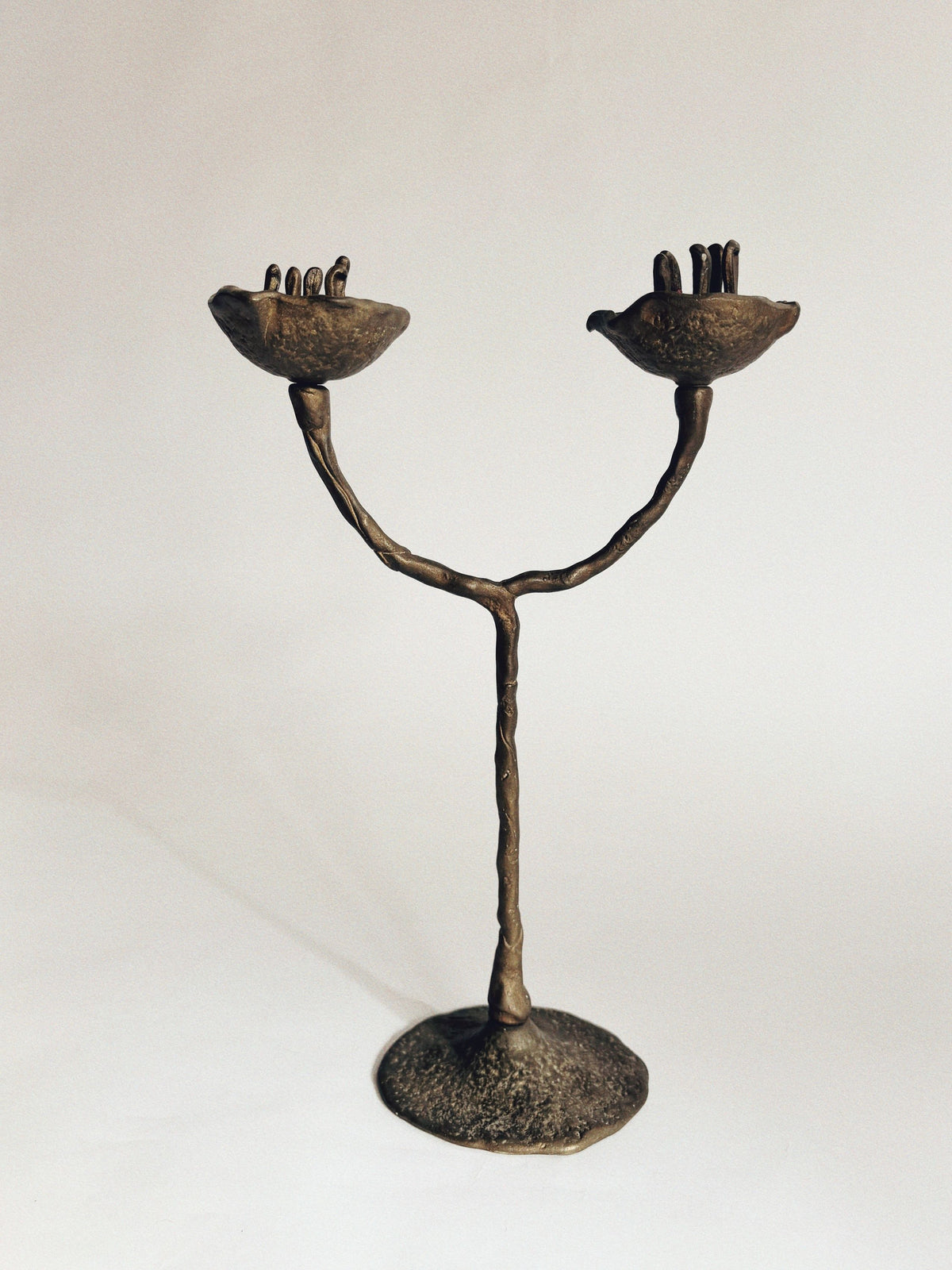 The Huerto 2 Arm Bronze Flower Candelabra, crafted in the 1990s by a Polish artist, features two curved arms ending in hand-shaped cups with upright fingers, all supported by a textured base and set against a plain background.