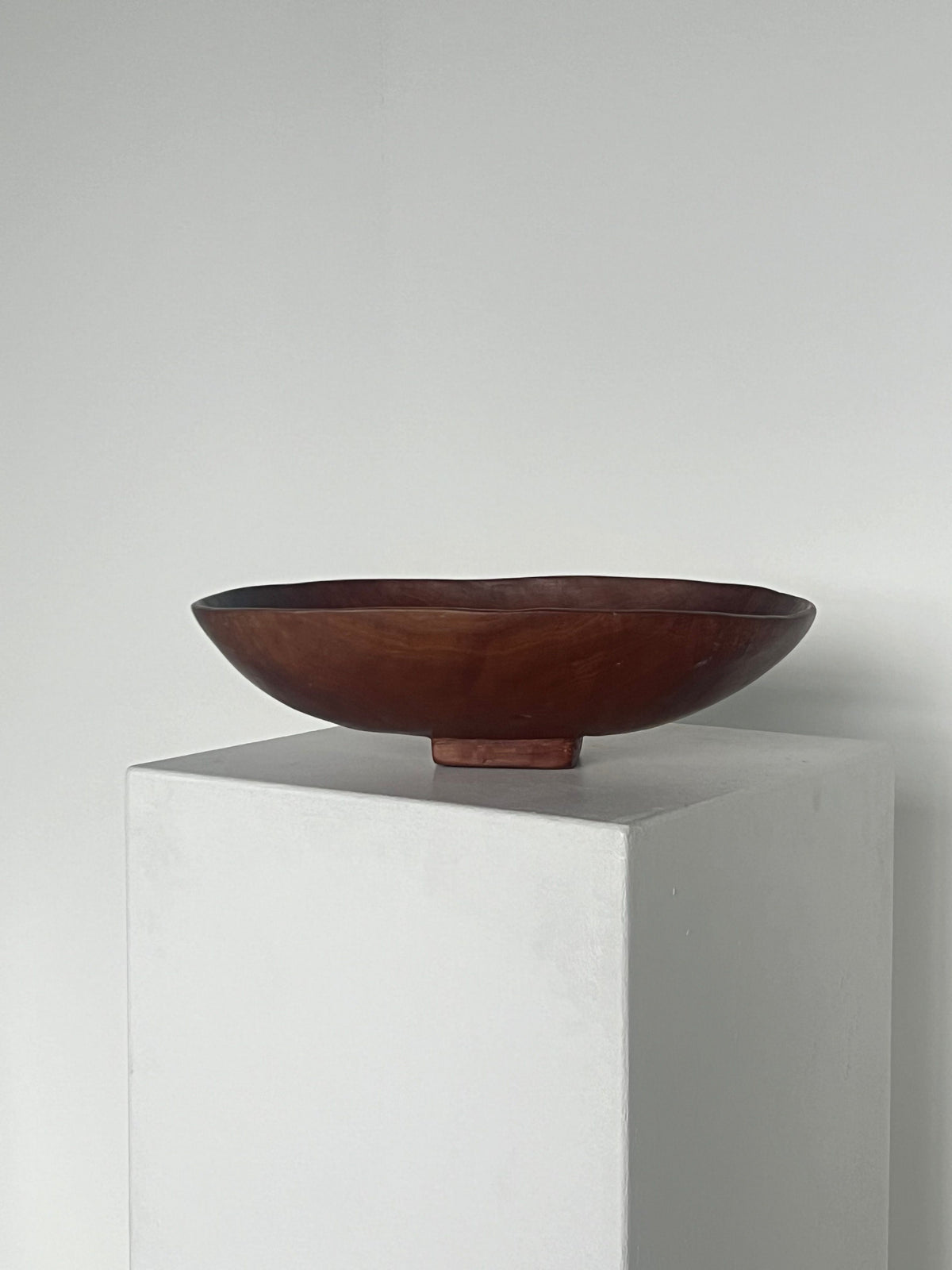 A Mahogany Hand Carved Oval Bowl from Porter Studio is placed on a white pedestal against a plain white background. The minimalist setting emphasizes the bowl's smooth texture and curved shape, highlighting its exceptional craftsmanship.