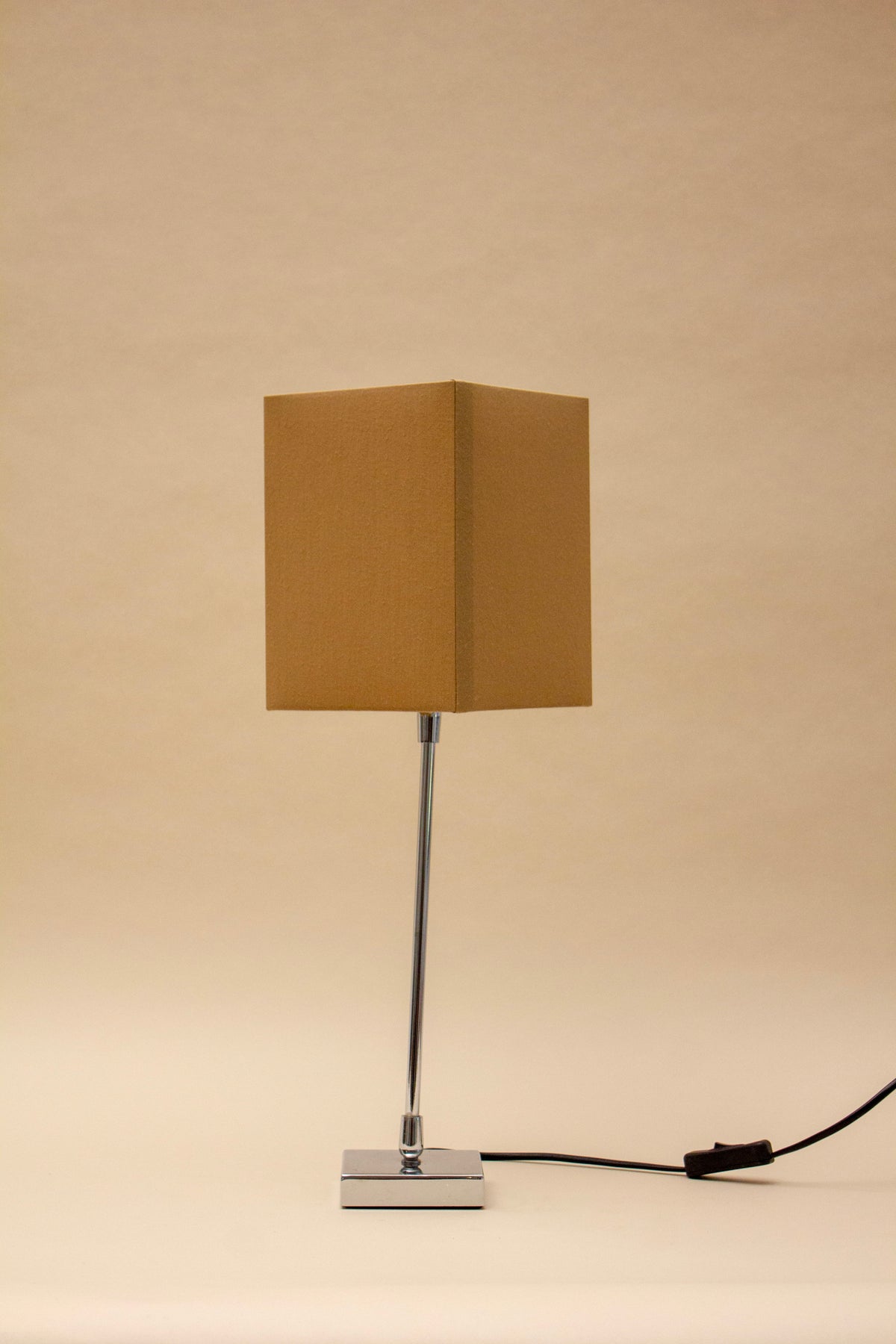 The ZAFRA 2 TABLE LAMP by SÒLER features a rectangular beige shade, slim metal stand, square metallic base, and a black power cord with switch on the right side, all set against a plain background.
