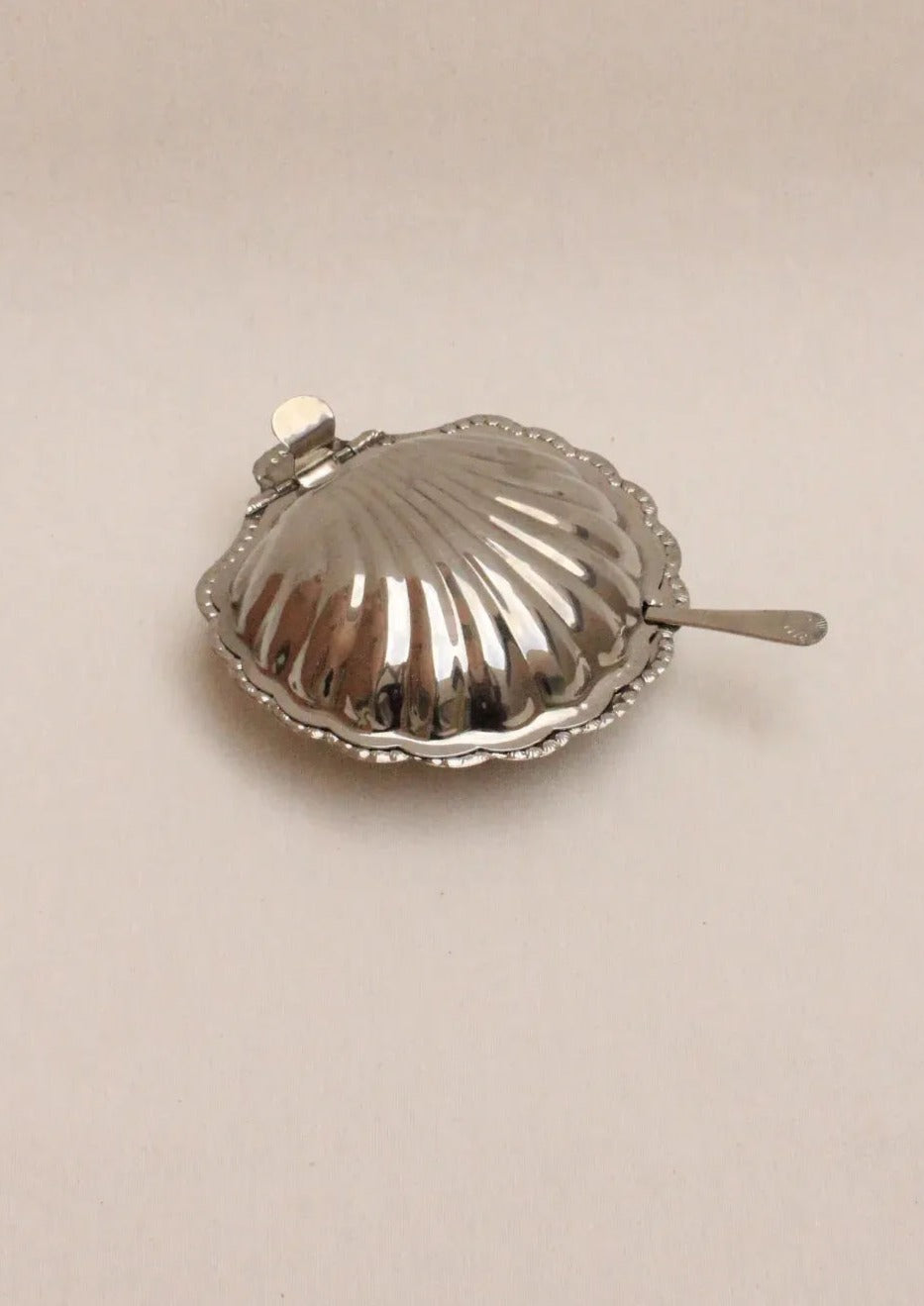 The Shell Shaped Butter Dish by Boga Avante Shop features ornate edges and a shiny silver finish, embodying a minimalist luxury aesthetic. It has a clam shape with a side lever, making it an elegant addition to any interior when placed on a light-colored surface.