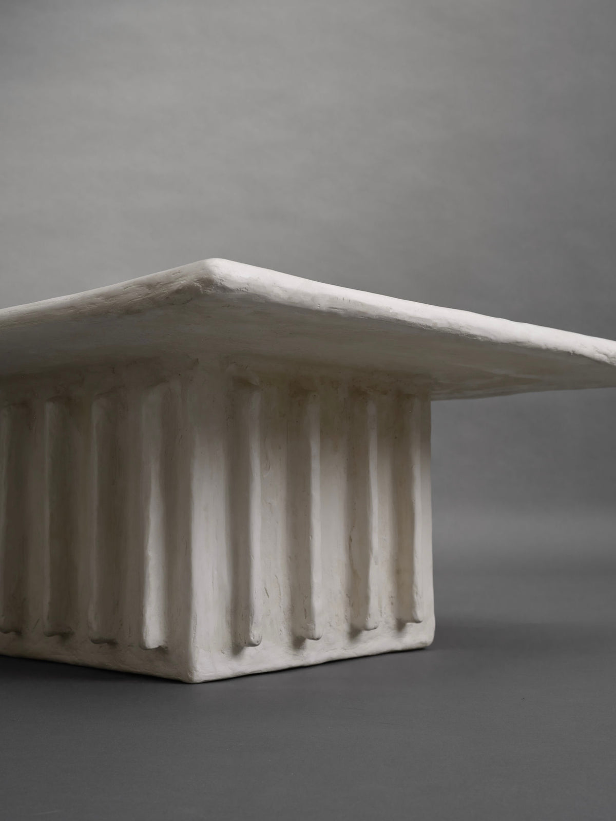 The Columnar Coffee Table by Ombia Studio is a minimalist white rectangular piece featuring a textured, ribbed base like classical columns. It's available in custom sizes against a smooth, muted gray background, highlighting its architectural design as a unique made-to-order item.