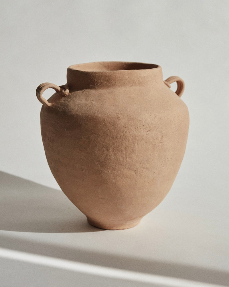 The Column Vase by marta bonilla, a rough-textured brown terracotta stoneware pot with a wide opening and two loop handles, rests on a white surface casting a shadow. Its rustic aesthetic is enhanced by the earthy tone, while the glazed inside adds an unexpected smoothness.