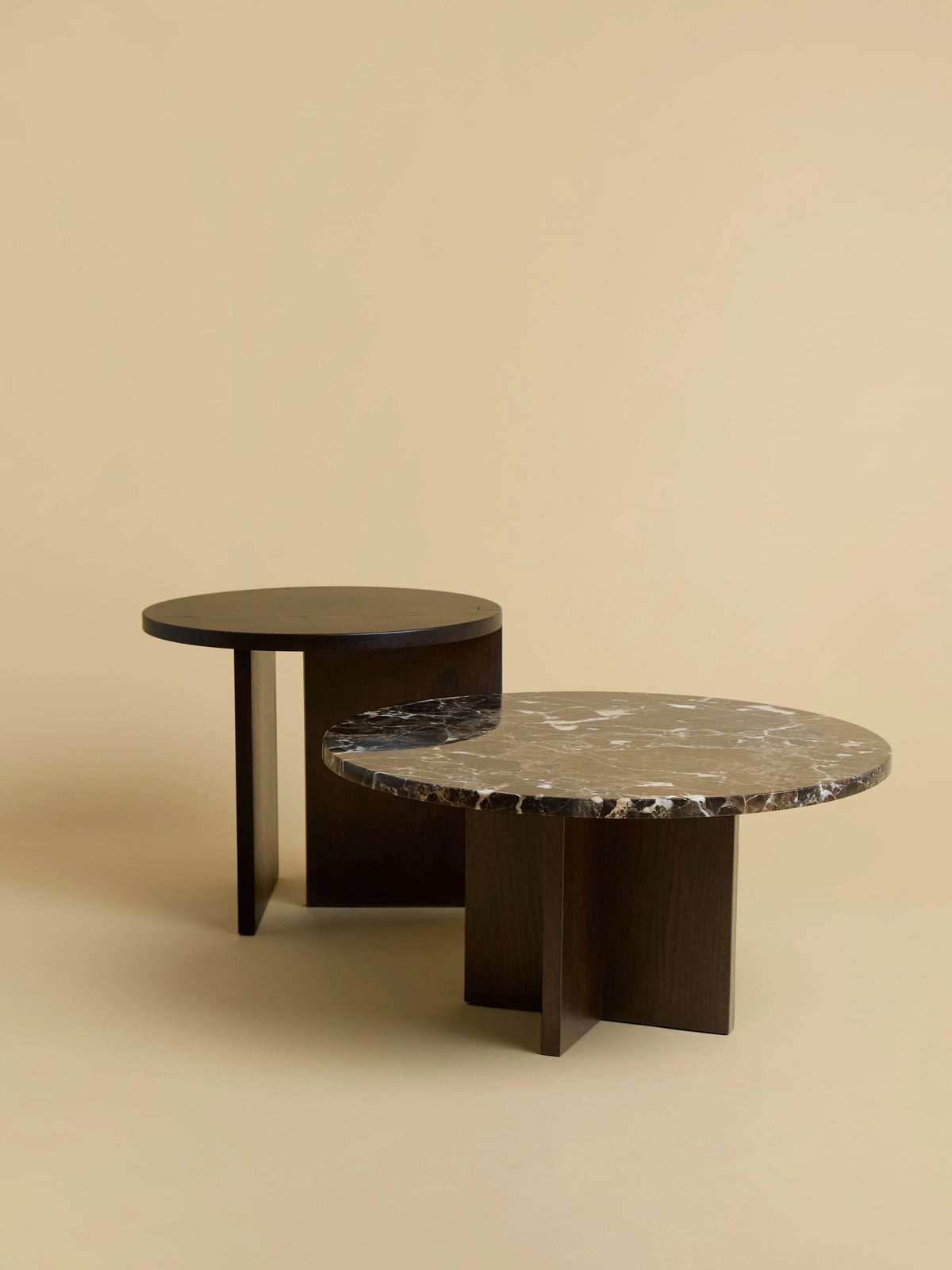 On a beige background, two round coffee tables are displayed. The front table features a dark wooden base with a black marble top and white veining, while the back table is slightly smaller and is "Kume" by Lebanto, made entirely of solid oak wood.