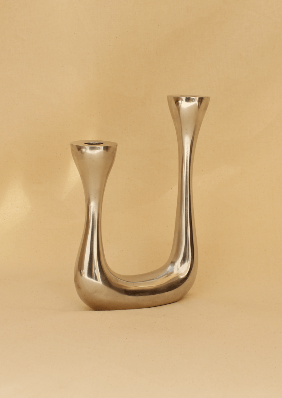 The Brutalist Candelabra by Boga Avante Shop is a modern, minimalist candle holder with a U-shaped metallic design featuring two handcrafted holders at different heights, set against a soft beige background.