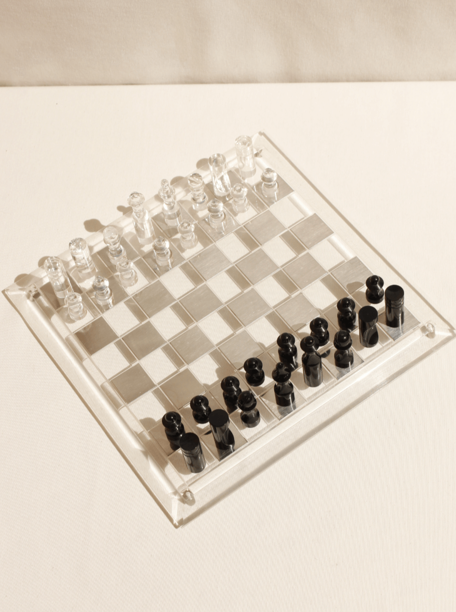 The Boga Avante Shop's Acrylic Chess Board features a sleek design with clear and black pieces. Pawns line the front, complemented by rooks, knights, bishops, queens, and kings in the back. This handcrafted set elegantly adorns any light surface for an intriguing game experience.