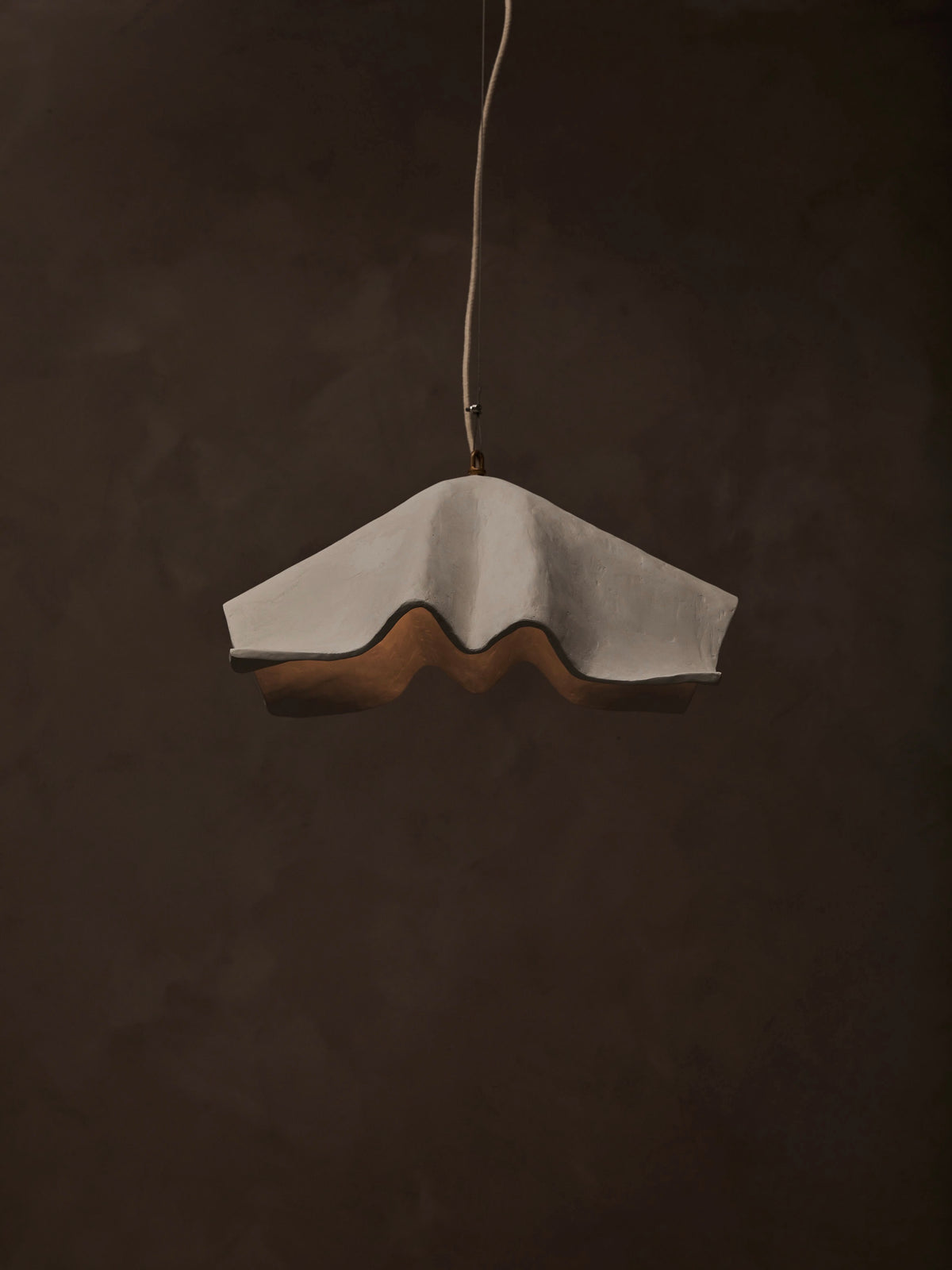 The 'La Fleur' Pendant Light by Studio Tristan Kallas, with a wavy umbrella-like design in white and brown shades, hangs from the ceiling against a dark background. It features dimmable lighting that casts subtle shadows on its smooth, textured surface.