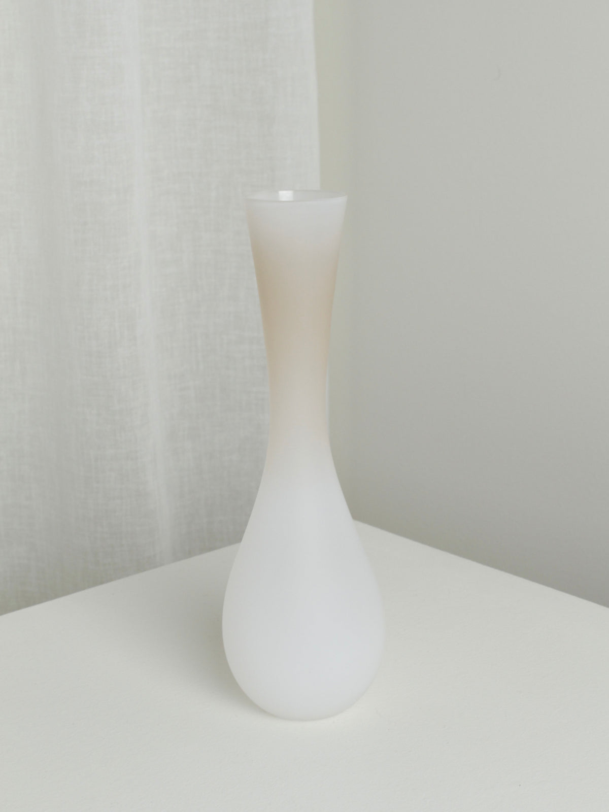 A minimalist Medium Vase in Mushroom by Verre d'Onge, featuring a frosted white finish with an elongated narrow neck and bulbous base reminiscent of its namesake, is placed on a white surface. The background showcases a light-colored fabric curtain and an off-white wall, creating a clean and serene ambiance.