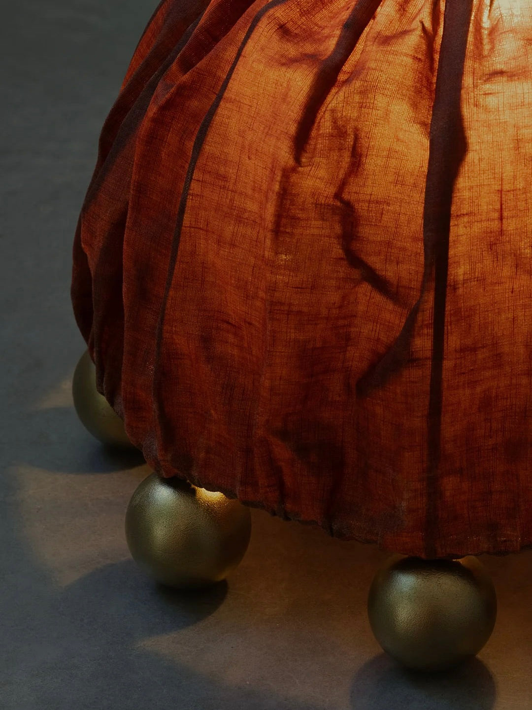 The ALTER Table Lamp by Kaytar features a crumpled, rust-orange beeswax-coated linen with vertical dark lines and alchemic symbols. It rests on five round brass feet, set on a dark surface, with lighting accentuating its intricate material and design.