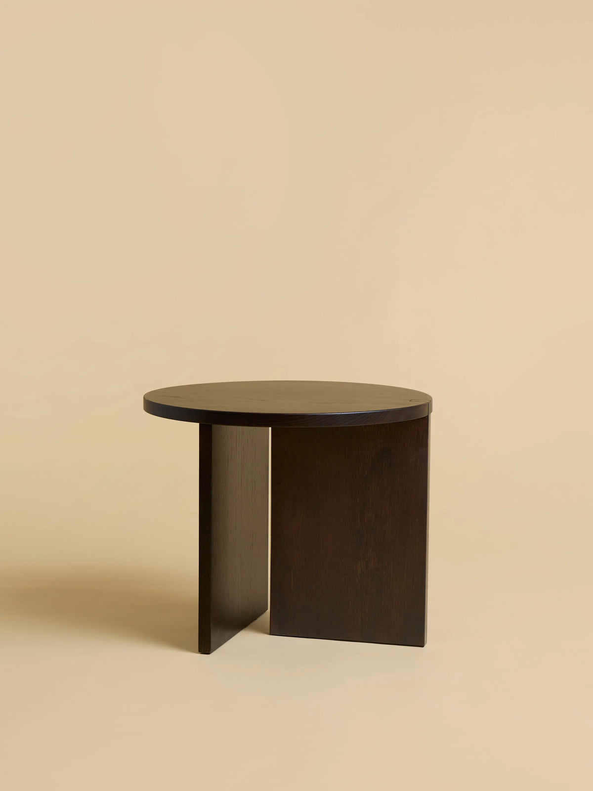 The "Kume" Side Table by Lebanto is a modern, round piece crafted from solid oak. Its versatile design features a circular top supported by two perpendicular rectangular panels with warm brown tones, complemented by a soft beige background.