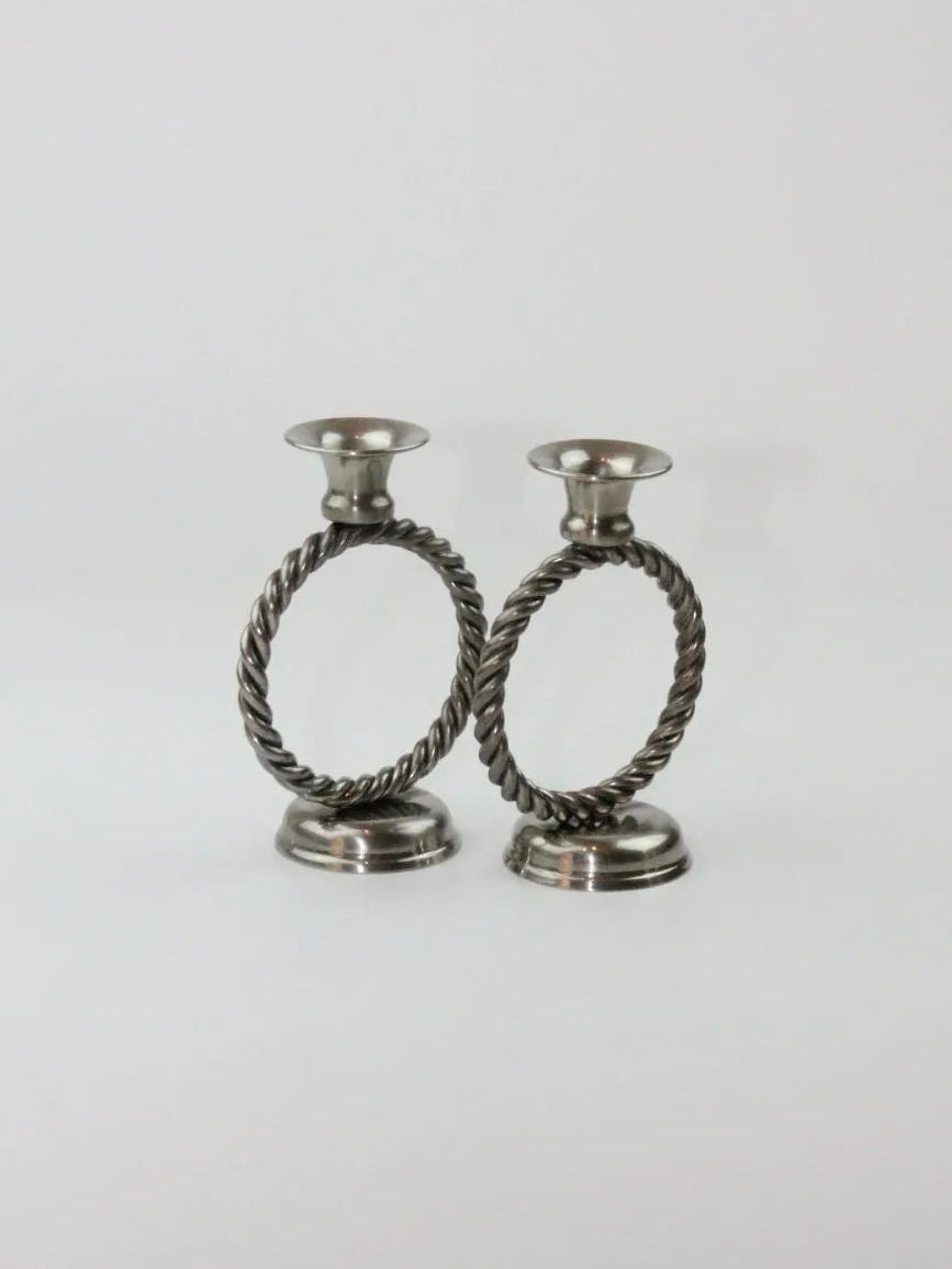 Introducing the Mental Candle Holders from Boga Avante Shop, a pair of classic and sophisticated silver candle holders featuring elegant twisted circular designs. Each metal holder is designed with a round base and a cup at the top for placing candles. Set against a plain, light-colored background, their timeless design truly stands out.