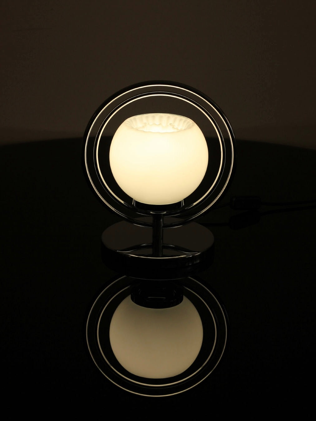 The Space Age Table Lamp from Boga Avante Shop features a modern spherical glass shade encased in concentric metal rings, all set on a reflective black surface. It emits a soft warm glow that creates a serene ambiance with its minimalist design.