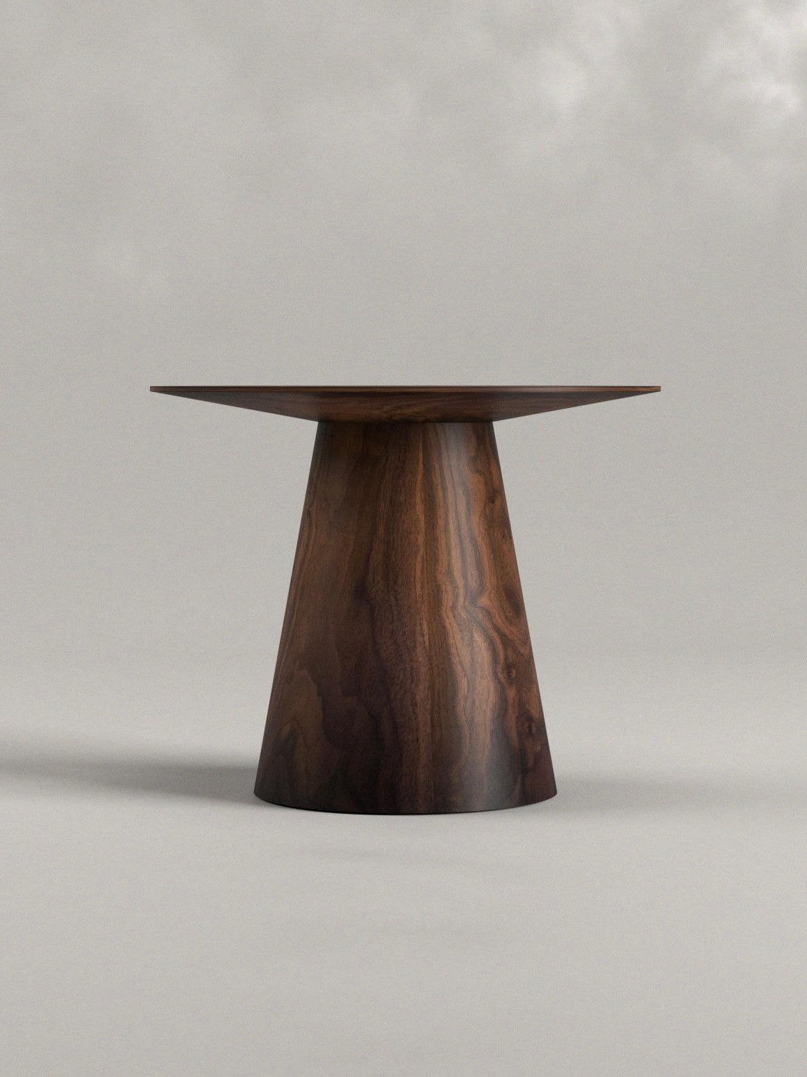 The Totem Side Table by Siete Studio is a minimalist piece with a sleek conical base and flat circular top. Handcrafted from walnut, its visible wood grain elegantly complements the soft gray background.