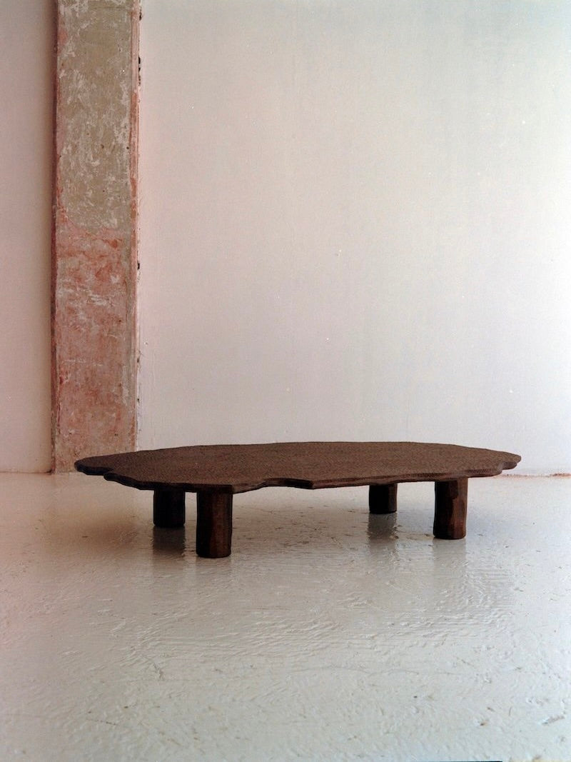 The Mare Coffee Table from Ombia Studio features a dark, organic-shaped solid wood top and four short legs, creating a striking centerpiece in a minimalist room with textured white walls and a weathered column.