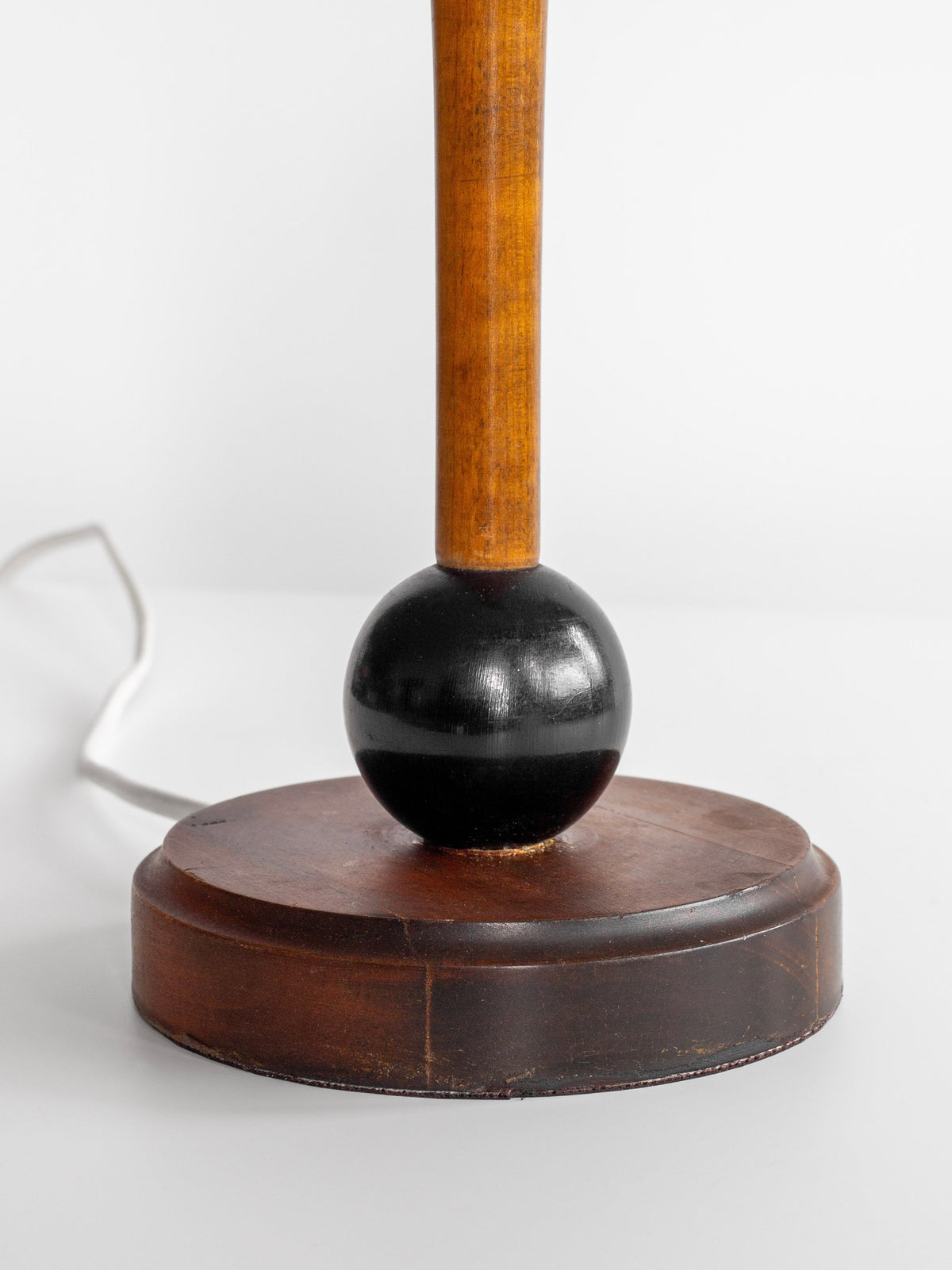 A close-up image of the Wooden Art Deco Table Lamp by Spigel. The round base is made of dark brown wood, featuring a spherical black element at the center that supports the wooden lamp stem. A white electrical cord extends from the base, evoking designs from 1930s Sweden.