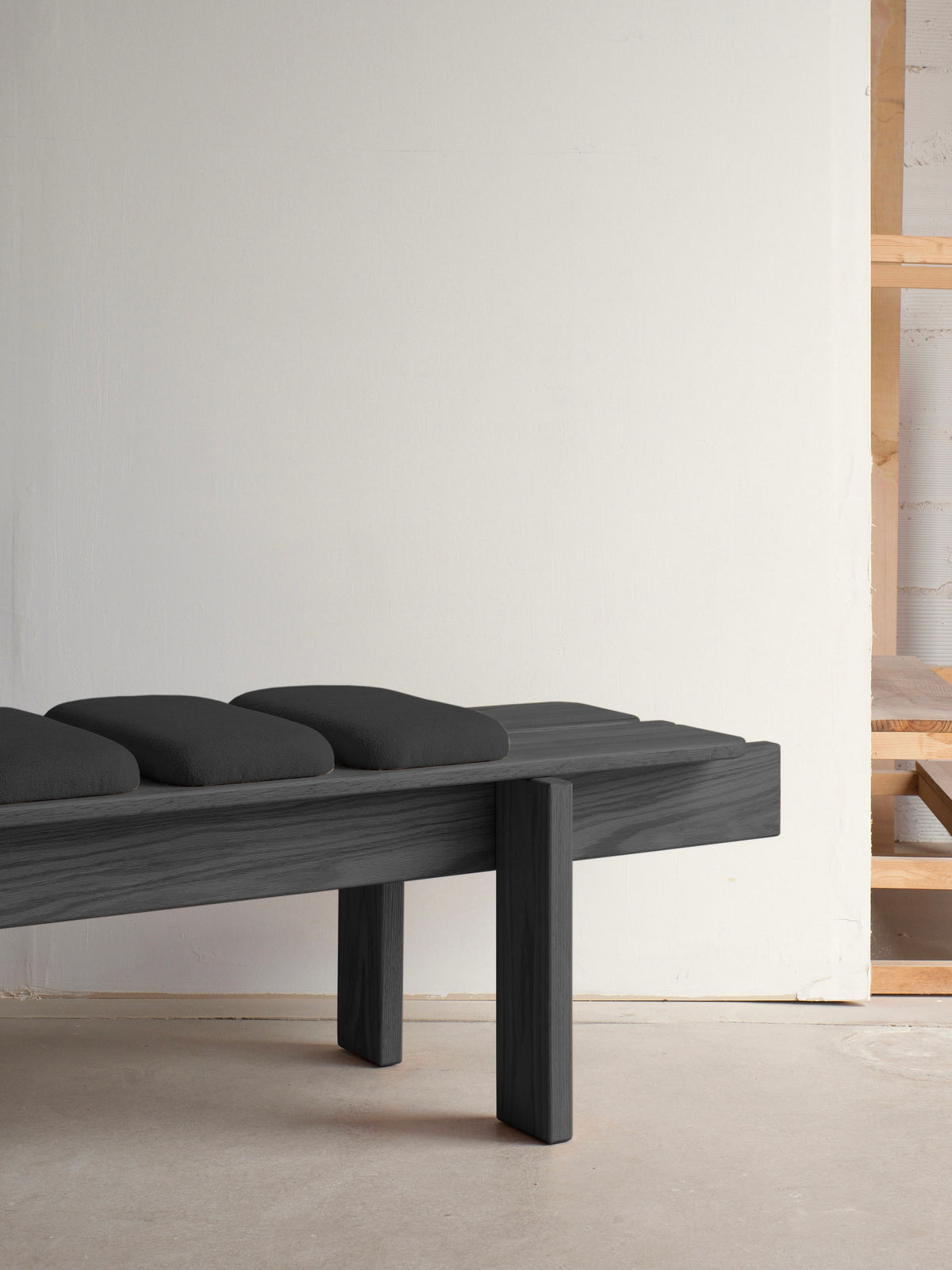 The minimalist scene spotlights the malva Ueno Bench Black Edition against a light wall, showcasing Japanese craftsmanship. Crafted from solid oak, the simple rectangular bench with sturdy legs and dark cushions stands on a bare floor. Wooden shelves in the background enhance the minimalist aesthetic.
