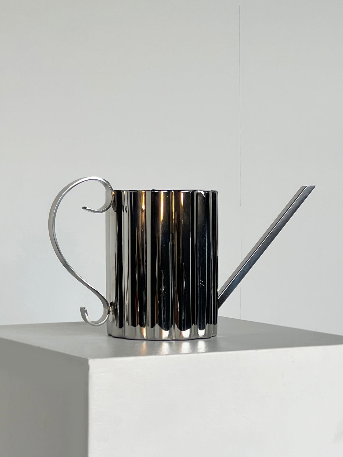 The Fluted Watering Can by Porter Studio, featuring a ribbed texture and long spout, is set on a white pedestal against a plain backdrop. Its silver-plated, reflective surface brings a modern flair to the minimalist composition.