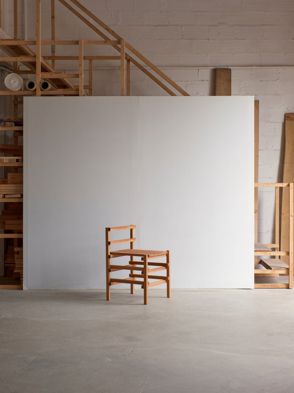 A minimalist Small Palma Chair by malva, featuring a ladder-like backrest, is positioned in front of a large white panel. The setting includes a wooden framework staircase that ascends to a loft, accompanied by various wooden materials and tools nearby, crafting a versatile statement piece within the workshop's ambiance.