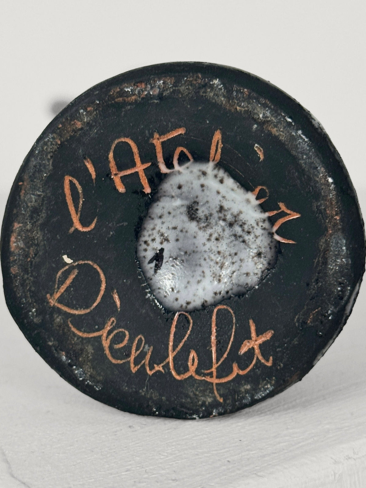Close-up of the Ceramic Bottle by Jacques Pouchain, from Figuresdesign. This round, rustic ceramic bottle showcases the hand-painted text "L'atelier Dieneslit" with a rough texture and a white glazed center. Reminiscent of Pouchain's classic style, it features an intricate bird decoration. The background is a neutral, light gray color.