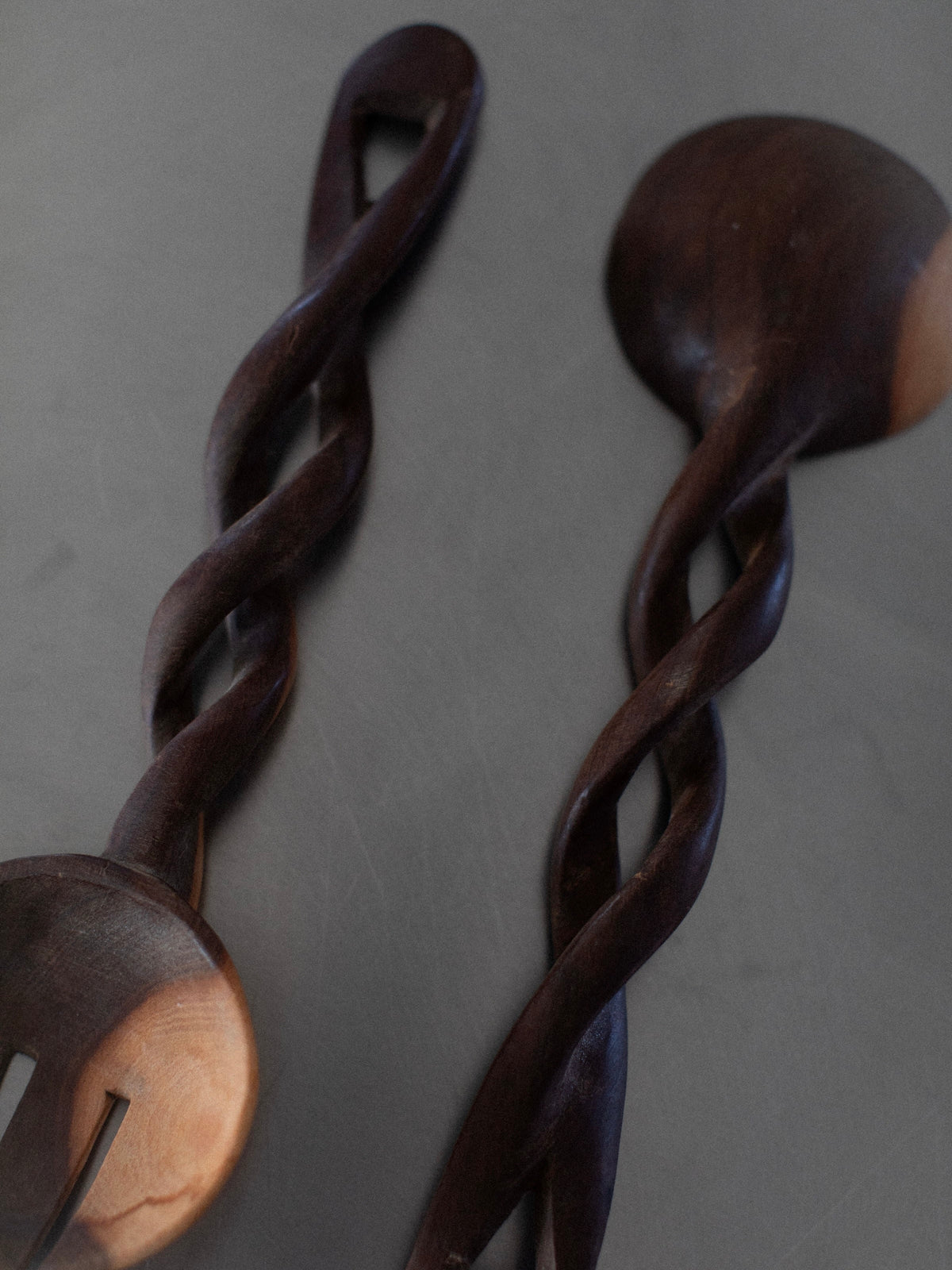 Two Twisted Serving Spoons by Out For Lunch, a spoon and a fork, laid parallel on a dark surface. Both handcrafted culinary tools feature intricately twisted handles, showcasing a unique and artistic design. The natural grain of the wood offers rich, dark tones that contrast beautifully with lighter patches on their heads.