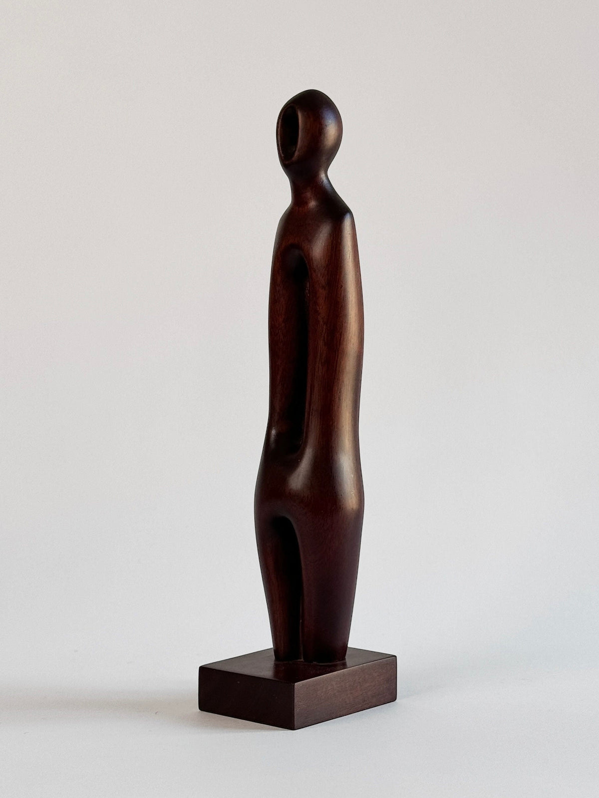 The Exotic Wood Sculpture by Pierre Dedieu from Figuresdesign stands on a small rectangular base. This unique piece features a smooth, dark wooden figure with a rounded head and elongated body, devoid of distinct facial features or limbs, giving it a minimalist and abstract appearance.