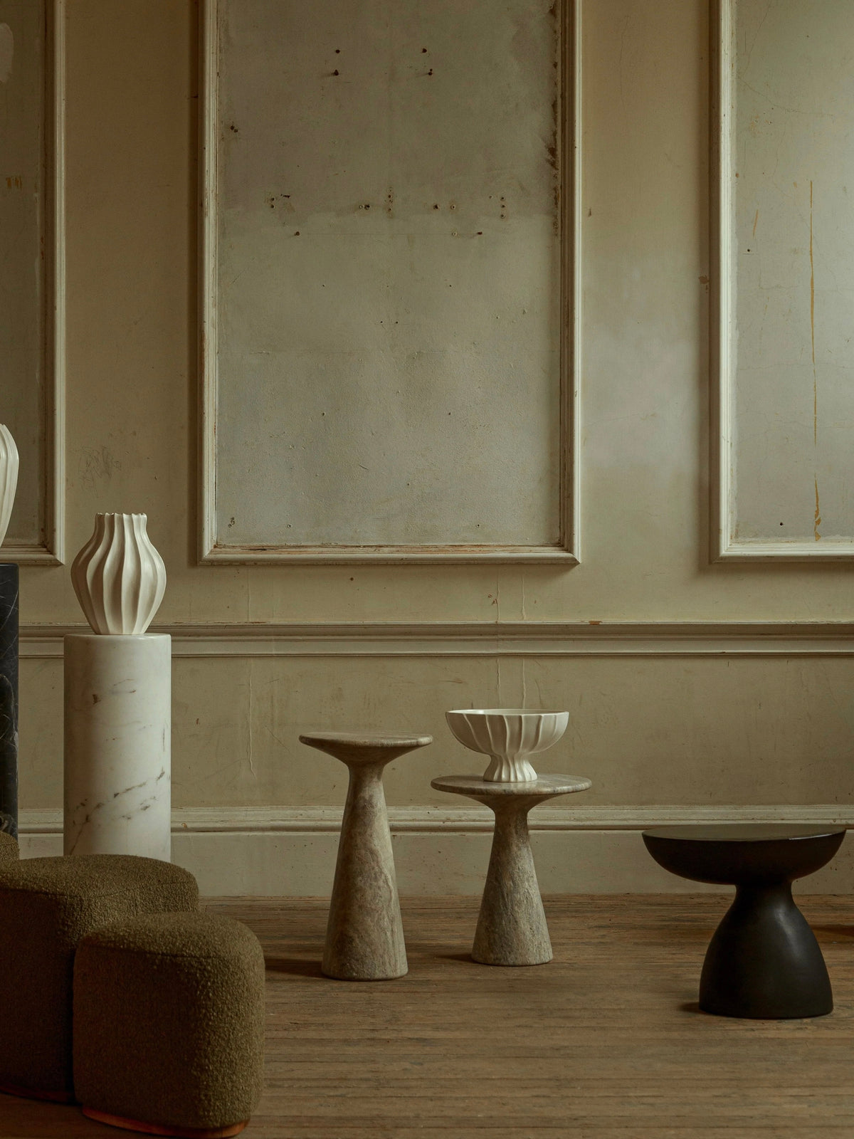 A minimalist room with textured beige walls and wooden flooring is adorned with abstract sculptures on marble pedestals, a green upholstered sofa, and intriguing stone tables. Among them is the "Cleo" Ceramic Side Table in matte black by Ransom & Dunn, which adds to the natural and artistic ambiance.