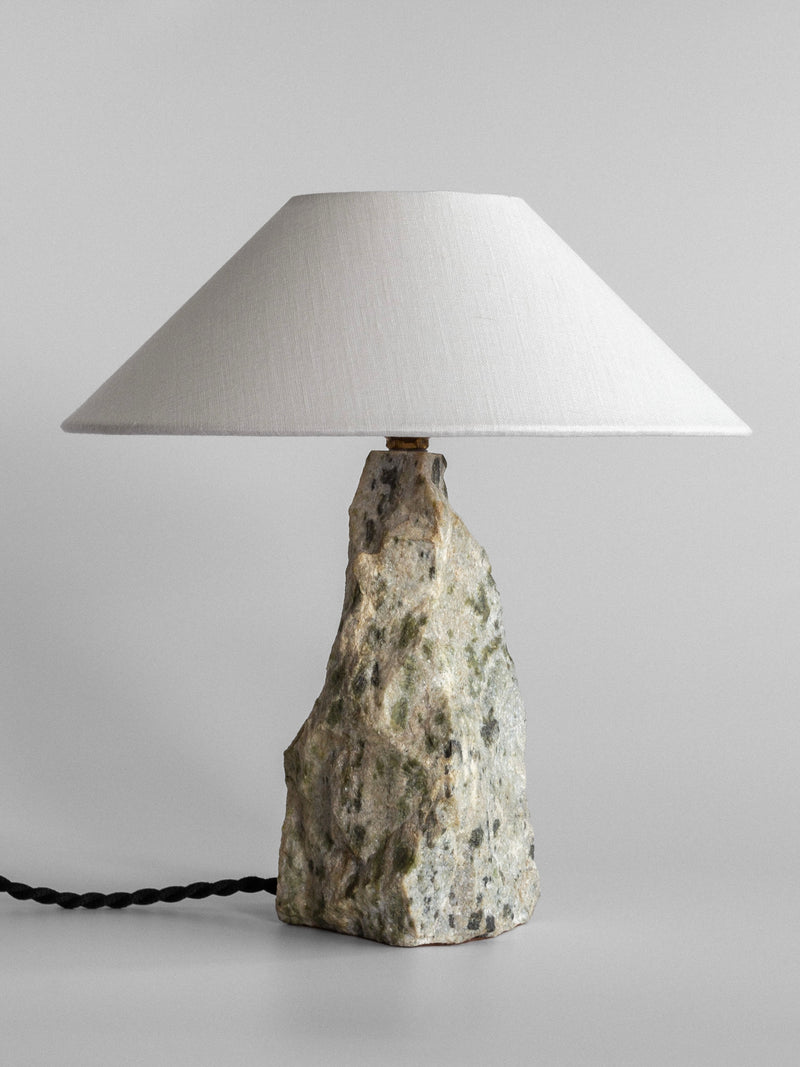 Introducing the Marble Stone Table Lamp by Spigel, featuring a striking jagged stone base inspired by Dunkeld marble and a broad, white fabric shade. Its sophisticated design is enhanced by a visible cord on the left side.