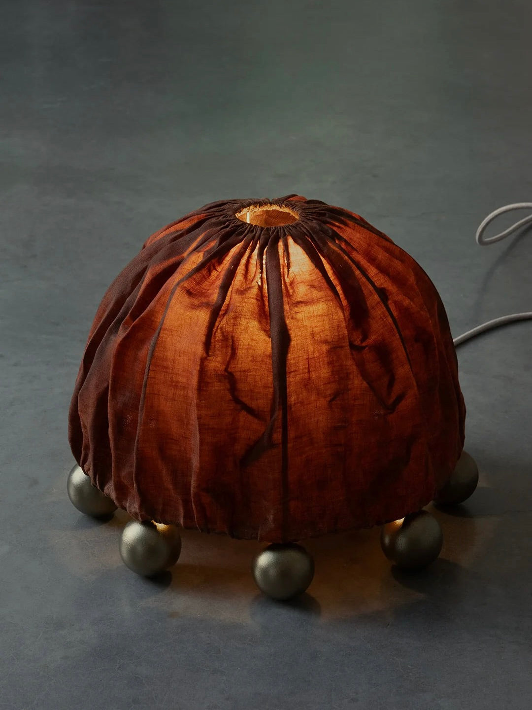The Kaytar ALTER Table Lamp features a wrinkled, beeswax-coated linen shade resembling a pumpkin. Resting on six small brass legs on a dark surface, this handcrafted lamp emits a warm, soft glow.
