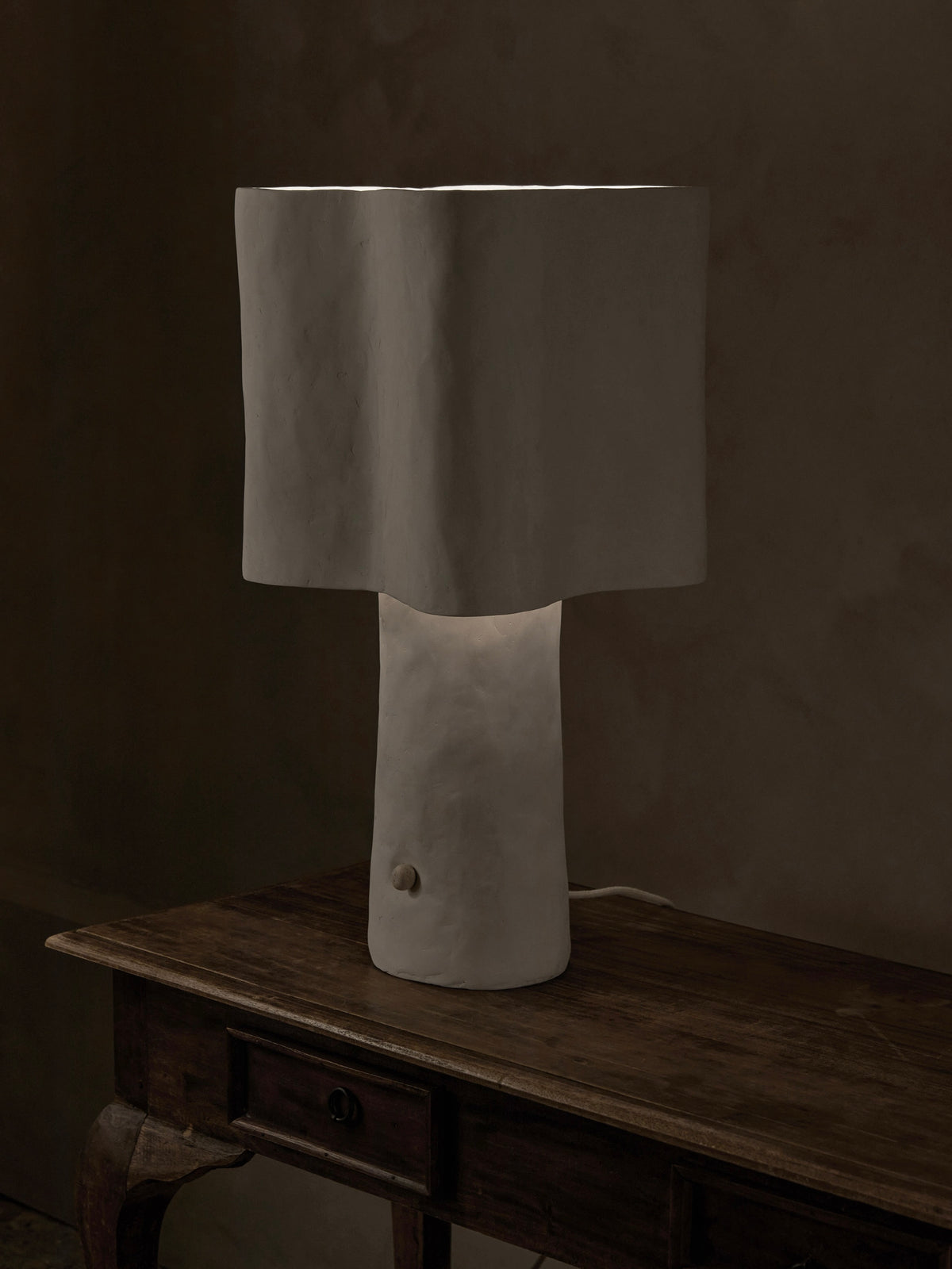 The Freeform Table Lamp by Studio Tristan Kallas is a minimalist sculpture with a handcrafted, textured white base and shade. Placed on a wooden table in dim light, it emits a warm glow that elegantly highlights the wood grain.