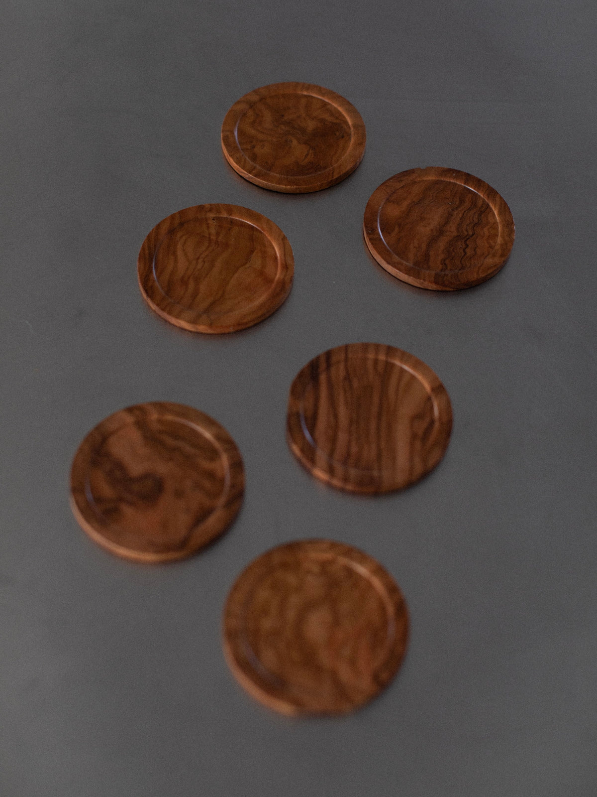 Six Out For Lunch Handcarved Coasters with visible grain patterns are arranged on a dark surface. The coasters exhibit intricate handcarved details and a smooth finish, showcasing natural wood tones from light to dark brown, offering a touch of natural elegance.