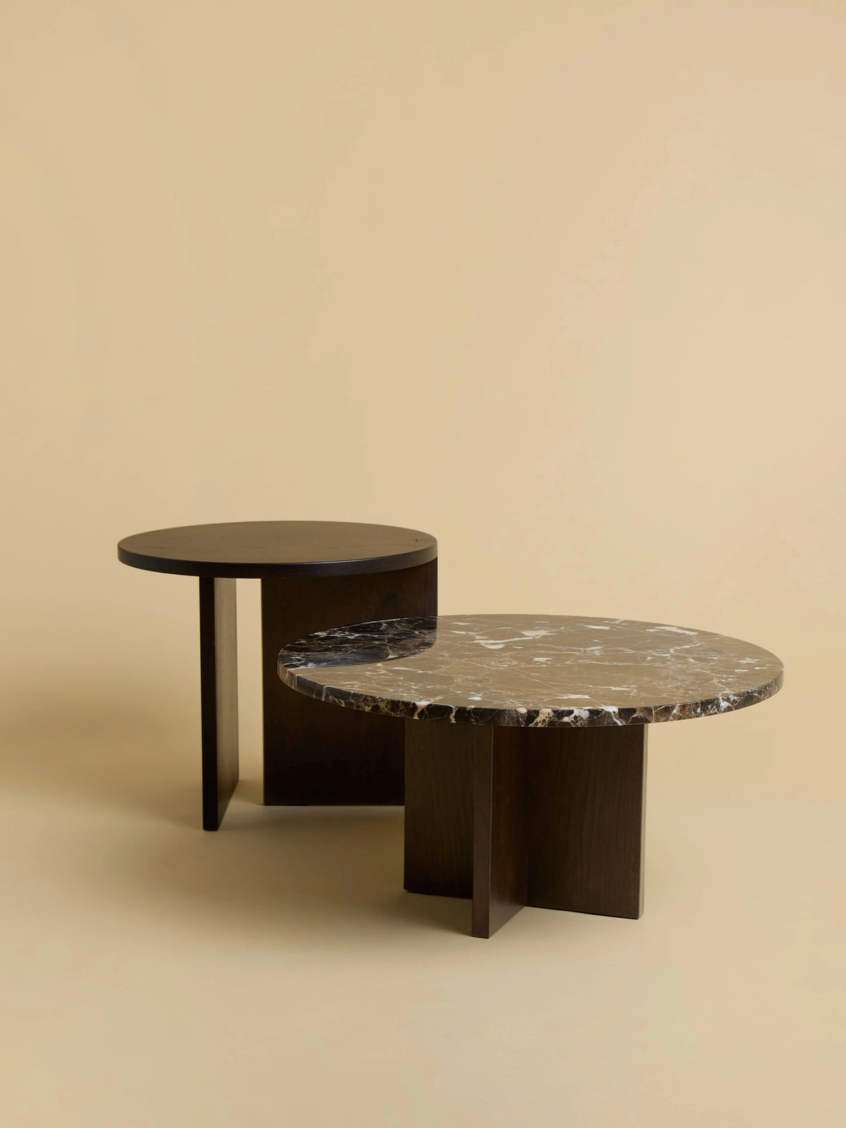 The "Tinian" Coffee Table set by Lebanto features two modern round tables with dark wooden bases. The front table has a marble top with white veining, and the rear table has a plain wooden top. Both are crafted with artisan precision and stand elegantly against a beige background.