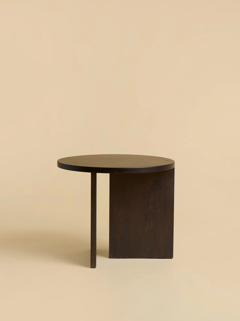 The "Kume" Side Table by Lebanto is a minimalist piece made from solid oak. It features an asymmetrical design with a dark finish, a circular top, one straight leg, and a wide curved panel, all set against a beige background.