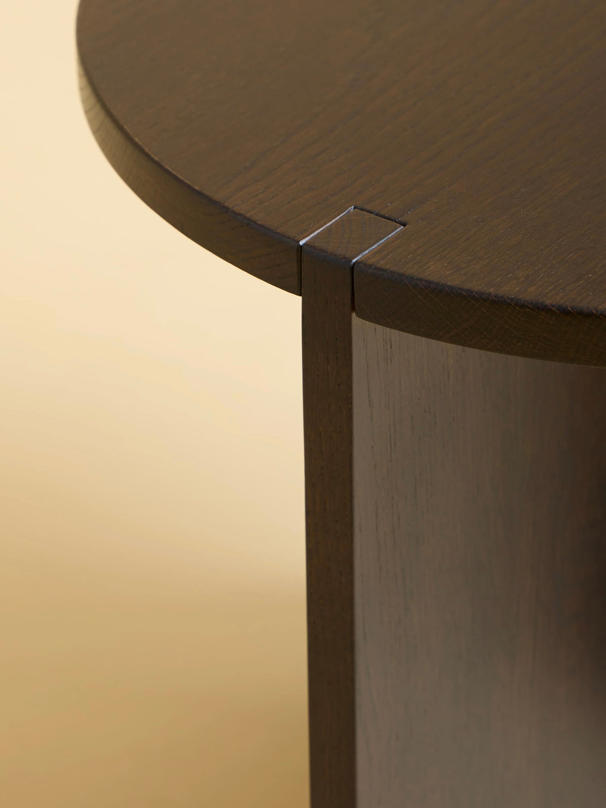 Close-up of the "Kume" Side Table by Lebanto, showcasing its smooth, dark solid oak wood and seamless joint connecting the round top to the leg. The soft beige background highlights its craftsmanship.