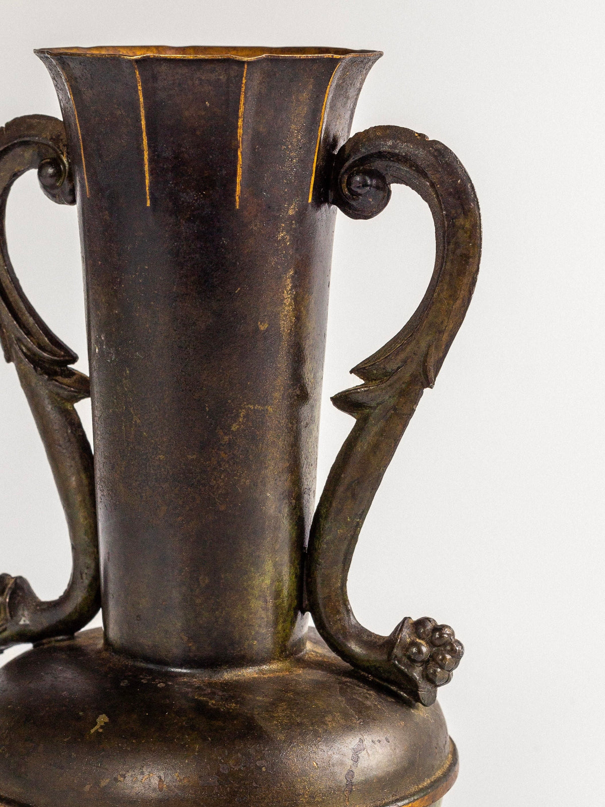 A close-up of the Spigel Art Deco Bronze Vase showcases its antique, dark metal finish with two ornate handles. The handles are gracefully curved with intricate detailing at the top and bottom, ending in a small cluster of rounded shapes. Resembling a neoclassical urn, the patinated bronze surface of this vase appears aged and weathered.