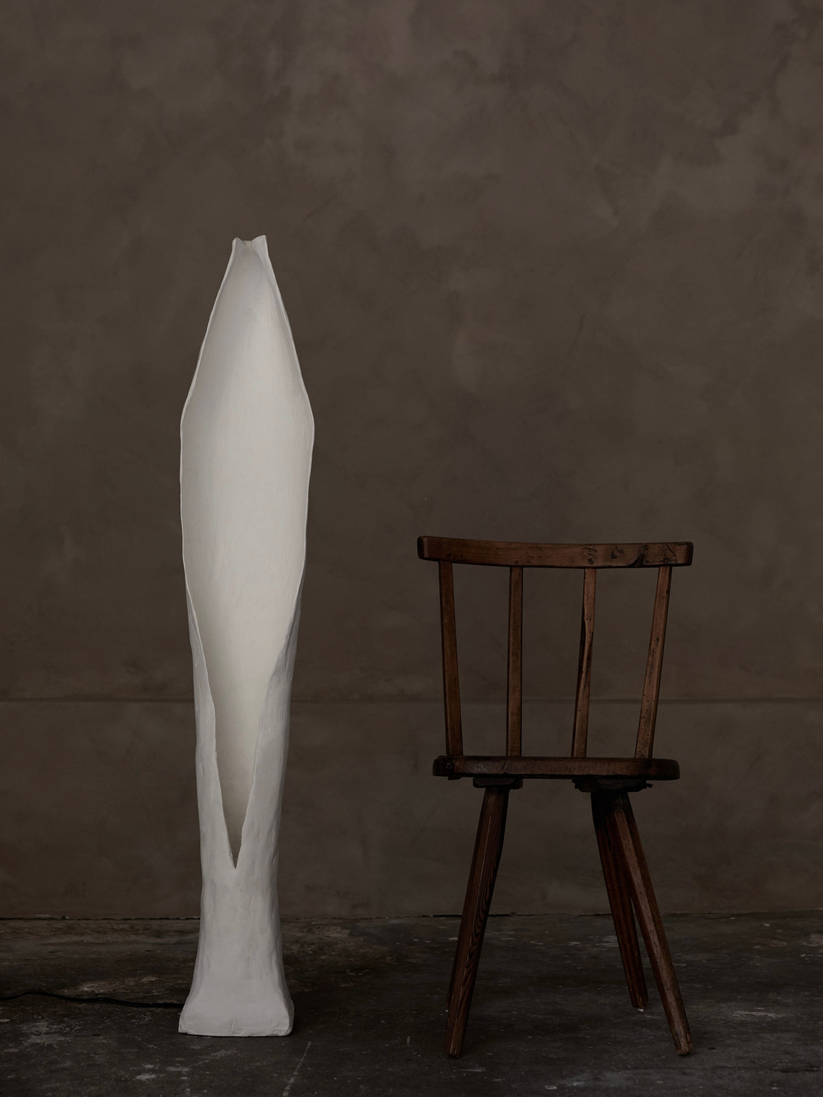 A tall abstract white sculpture like an elongated leaf stands next to a dark brown simple wooden chair, illuminated by the Alcove Floor Lamp. This serene arrangement by Studio Tristan Kallas is set against a textured taupe backdrop.
