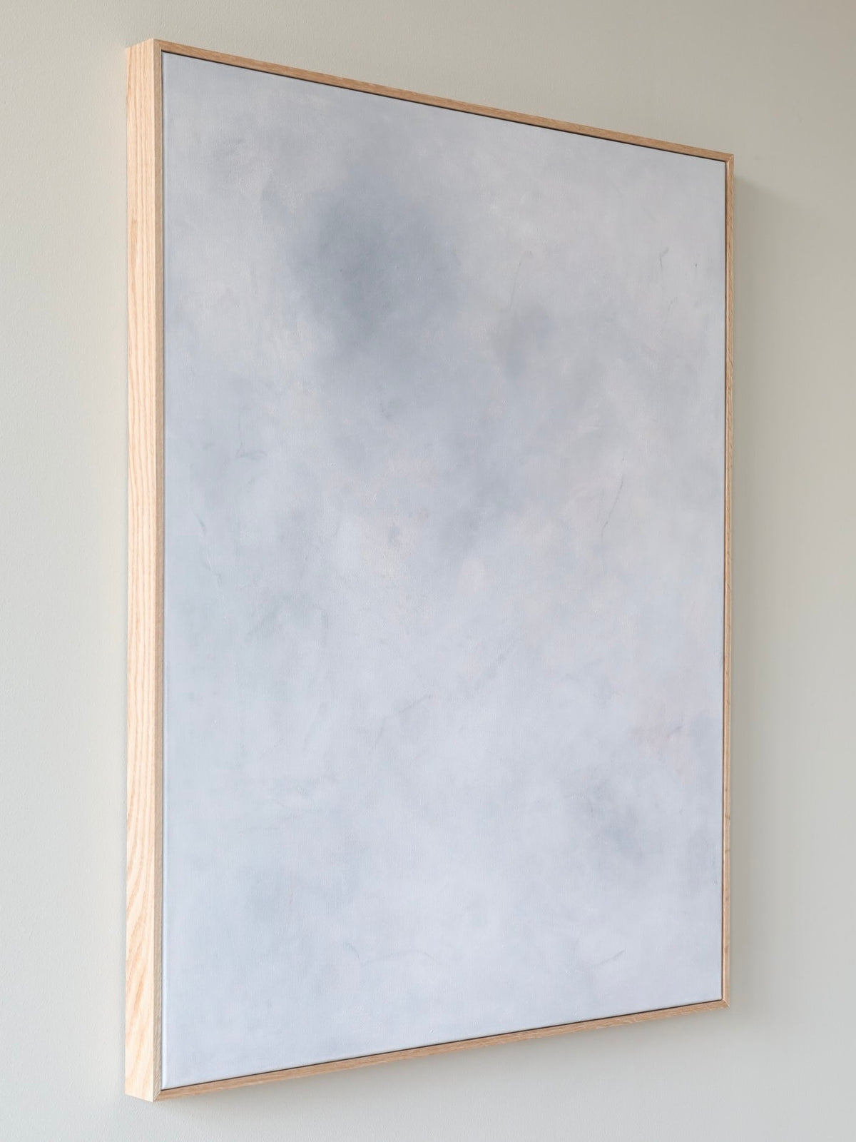 The Eos, Acrylic on canvas, 2024 by A Nesting Place is a minimalist acrylic painting featuring a light gray and white textured surface, framed in a thin, natural oak frame. This abstract canvas art showcases subtle variations in shading with faint hints of blue and is beautifully displayed against a pale wall.