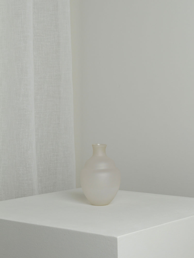 A Verre d'Onge Small Vase in Moon sits on a white pedestal against a backdrop of a white curtain, reminiscent of the calm night skies over Montreal. The scene is minimalist, with soft lighting that highlights the vase's smooth, rounded shape and the subtle texture of the curtain.