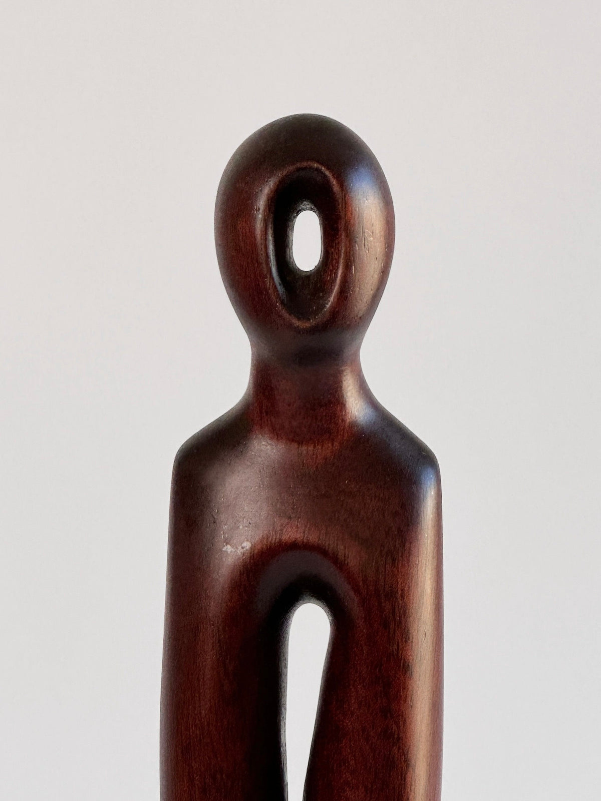 The Exotic Wood Sculpture by Pierre Dedieu from Figuresdesign is a minimalist wooden human figure featuring smooth, curved surfaces and clean lines. The figure boasts a rounded head with a hollow circular opening in the center and a vertical cut-out running from the torso to the lower body. This unique piece is polished to a dark finish using exotic wood.