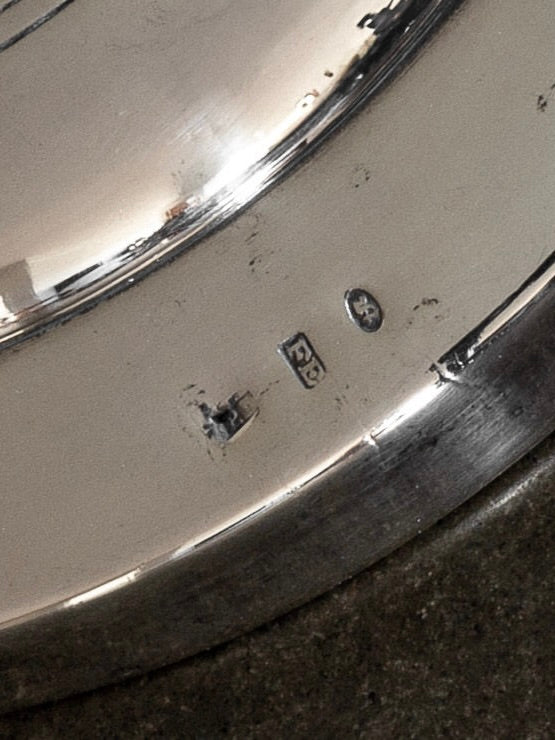 Close-up photograph of a silver-plated surface on the Spigel Silver-Plated Table Lamp, Early 1900s, featuring hallmarks or maker's marks reminiscent of Art Deco style. The slightly worn and scratched surface shows three distinct marks: one resembling a letter or symbol, another as a square with a design, and the third as a circular mark.