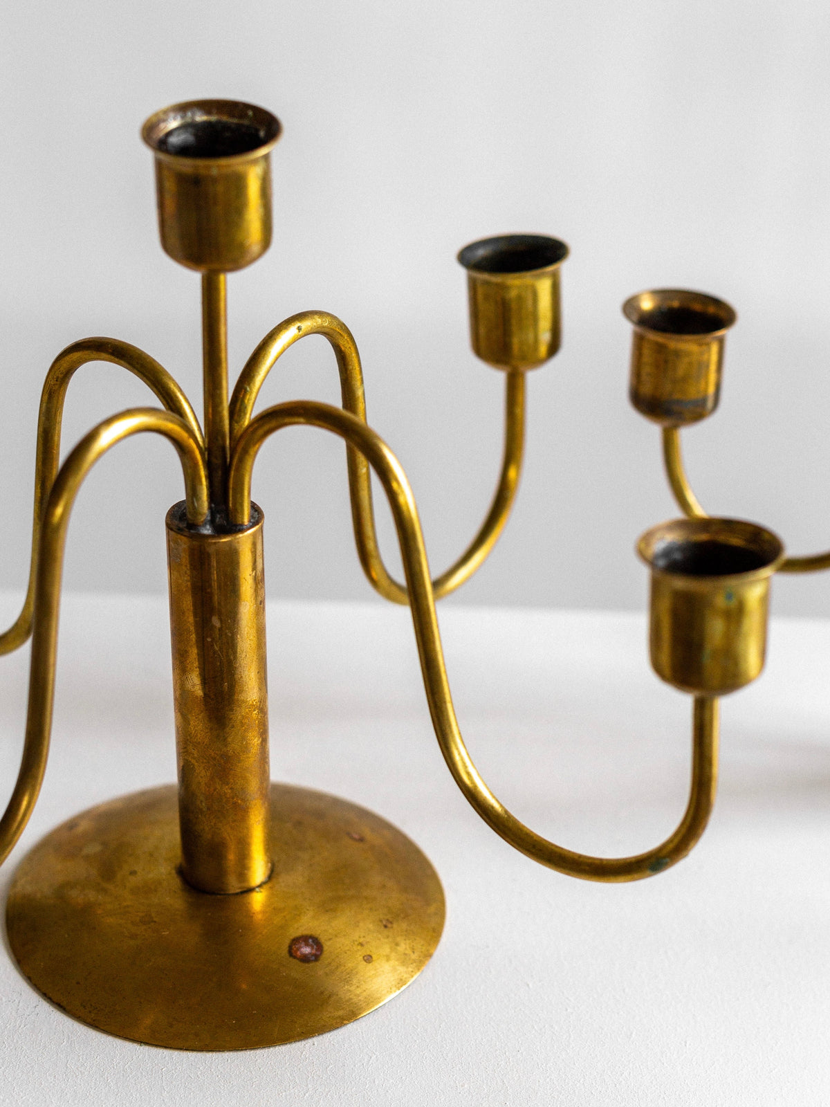 A Spigel Mid-Century Brass Candelabra with a central stem and five curved arms, each holding a candle cup. The candelabrum has a round base and is placed on a white surface. Its ornamental design exudes vintage appeal, and the brass has developed a slightly tarnished patina, enhancing its antique charm.