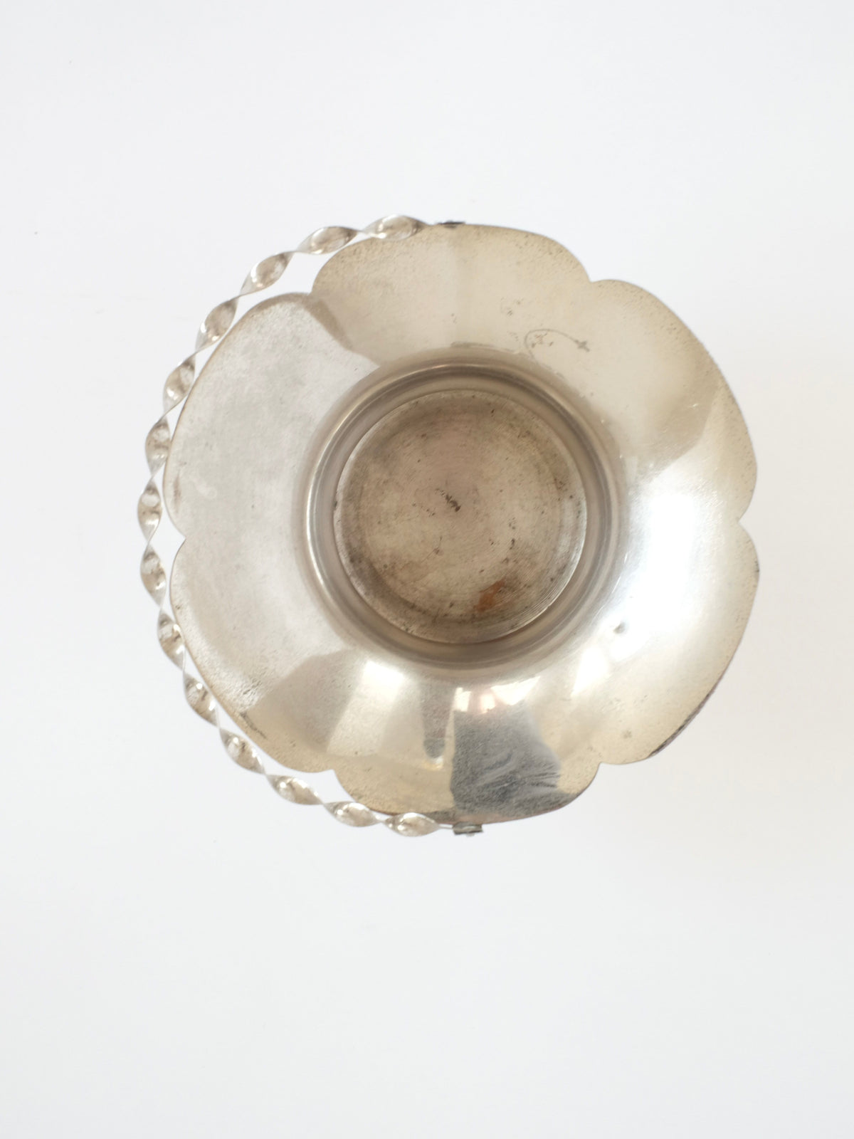 A top view of the "Basket With Twisted Hand Barrel" from septembre studios reveals a silver metal piece featuring a scalloped edge and a handle crafted from alternating shiny and matte metal orbs. This vintage-inspired item sits empty against a plain white background, reminiscent of an elegant silver basket.