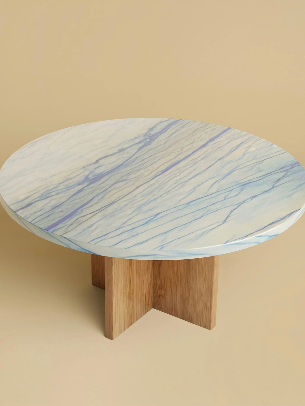 The "Tinian" Coffee Table by Lebanto features a marble top with subtle blue and gray veins, showcasing artisan craftsmanship. It has an oak wood base with four intersecting legs set against a beige background.
