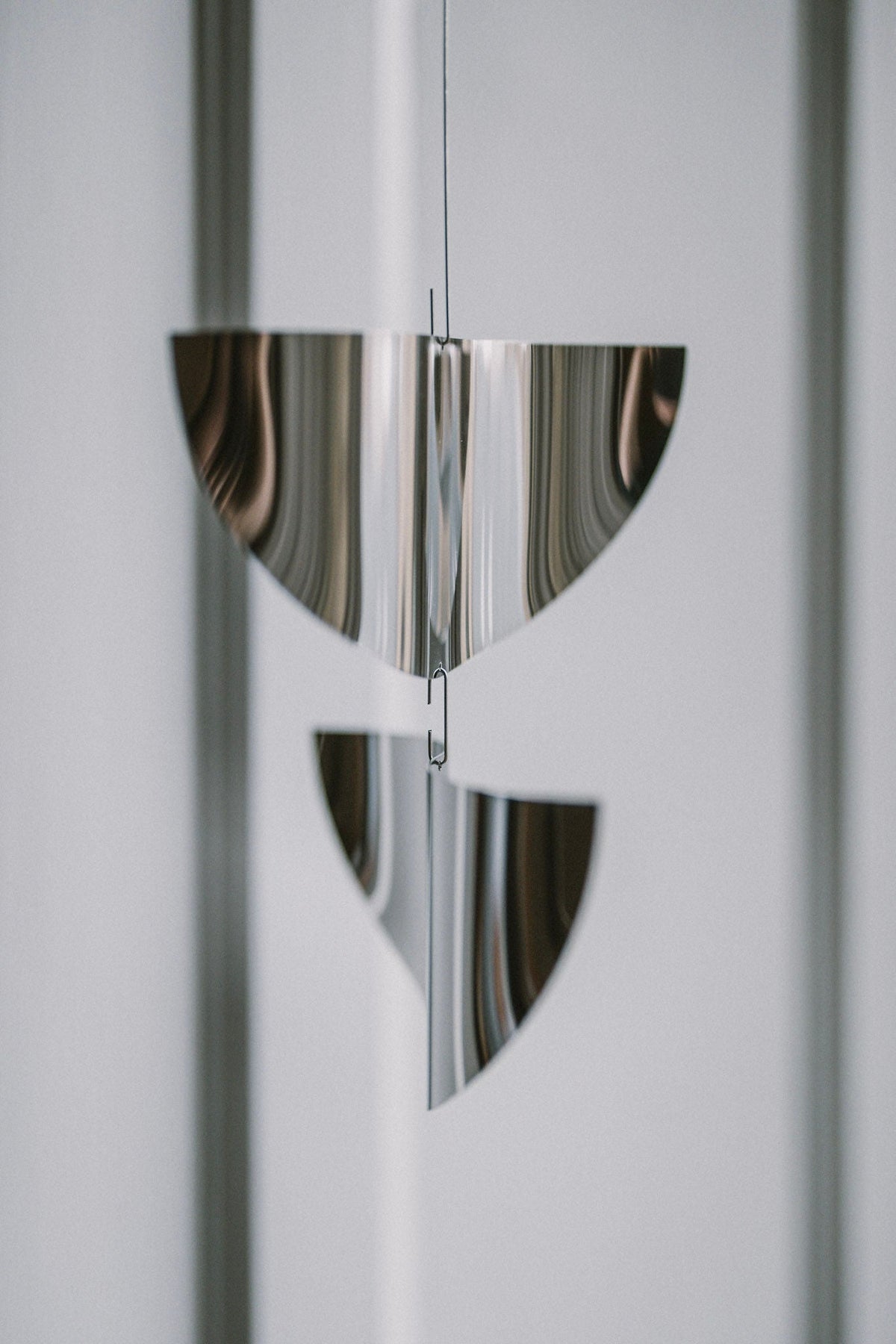 The Sun Catcher by Studio Fie Paarup is a handmade Danish metal mobile with two shiny, semi-circular forms suspended vertically. Its polished steel reflects light beautifully, adding a modern, abstract touch against a blurred gray background.