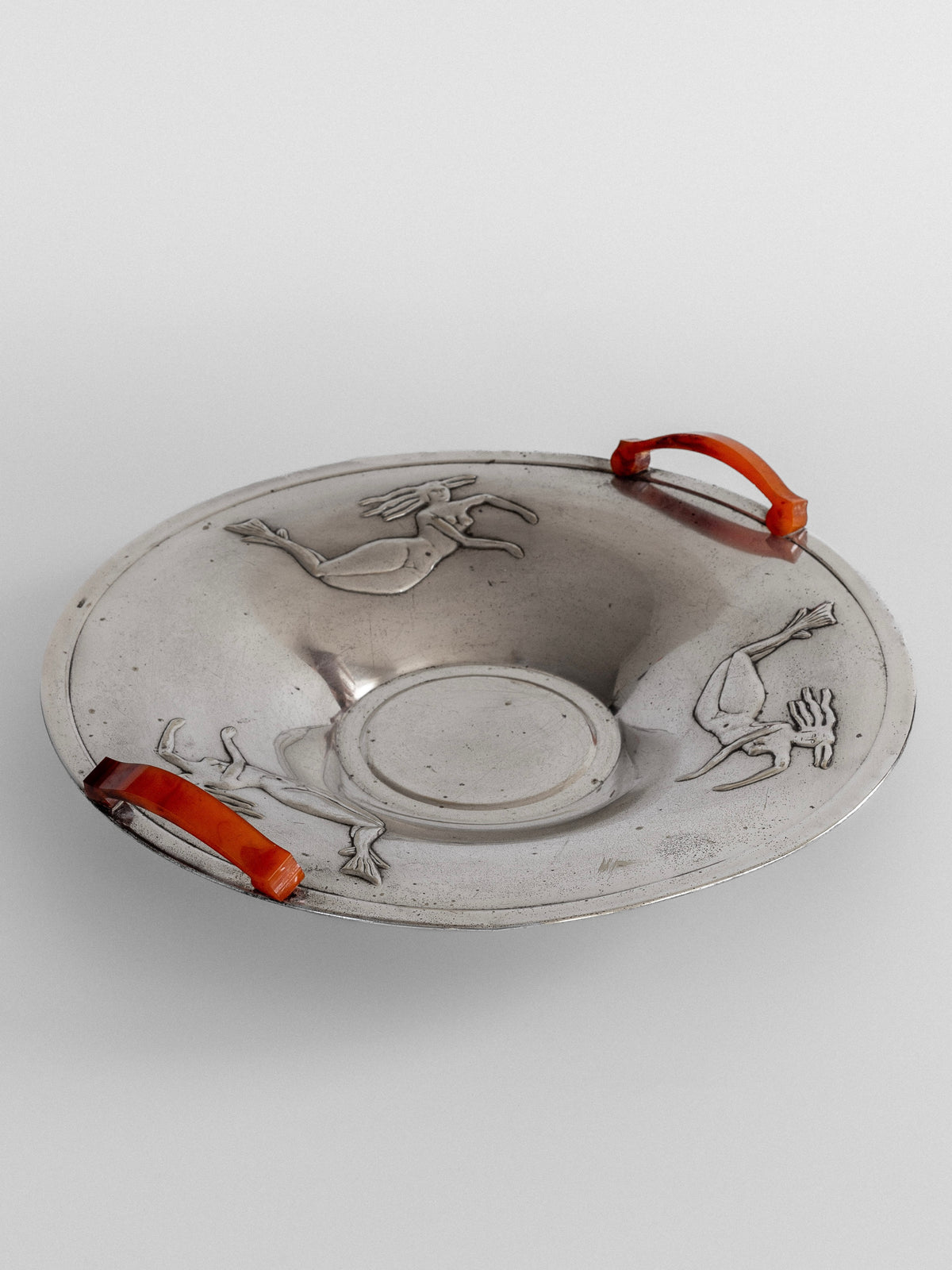 Introducing Spigel's Decorative Bowl with Mermaids from the 1930s: a silver-plated masterpiece adorned with two vibrant orange handles on opposite sides. This bowl, reminiscent of the Swedish Grace design, showcases engraved images of two figures in motion and features a central circular indentation.